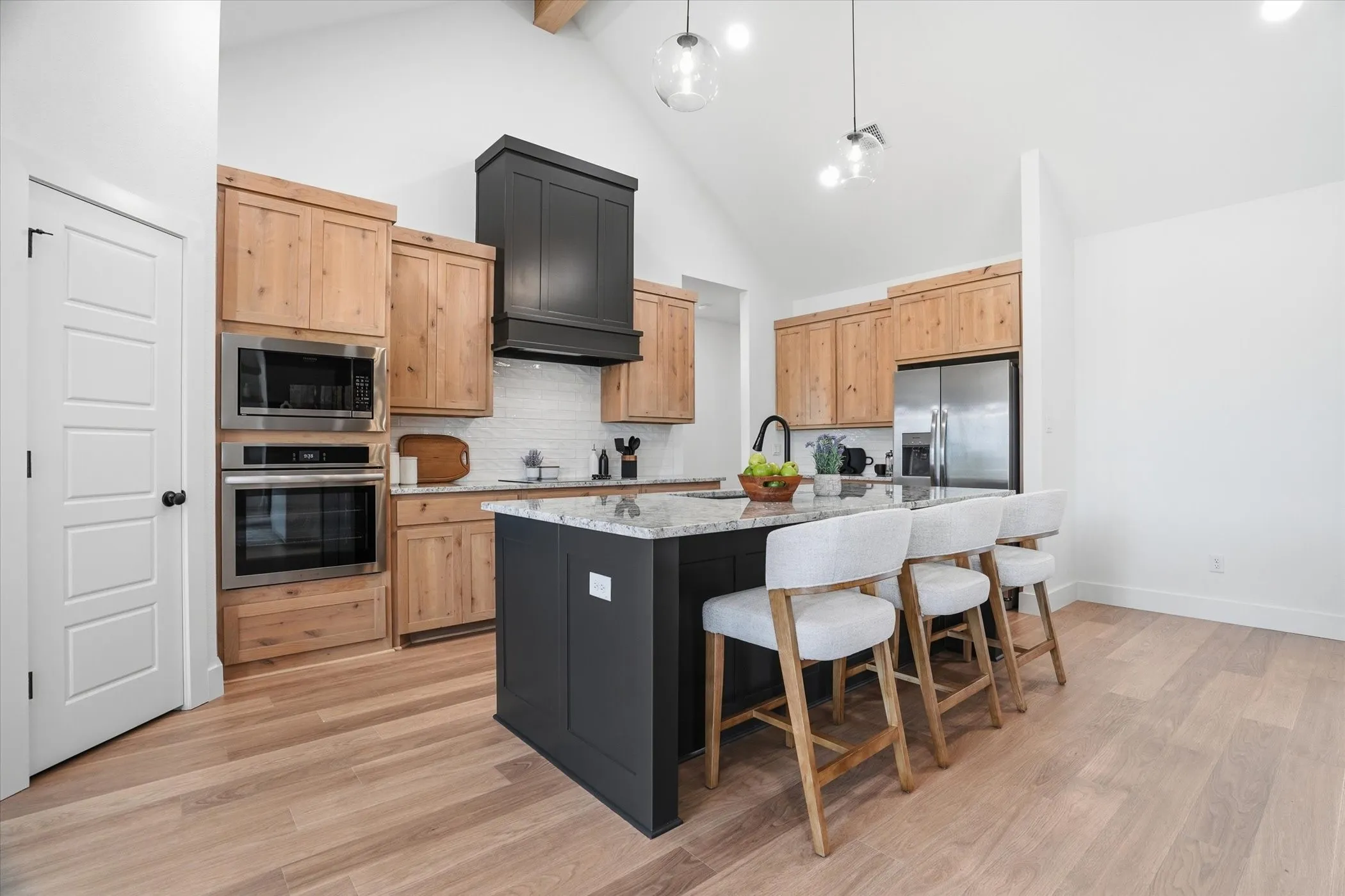 Vaulted ceiling with exposed beam, wood-finish flooring, built-in stainless steel oven and microwave, dark wood range hood, and a kitchen island with stone countertop