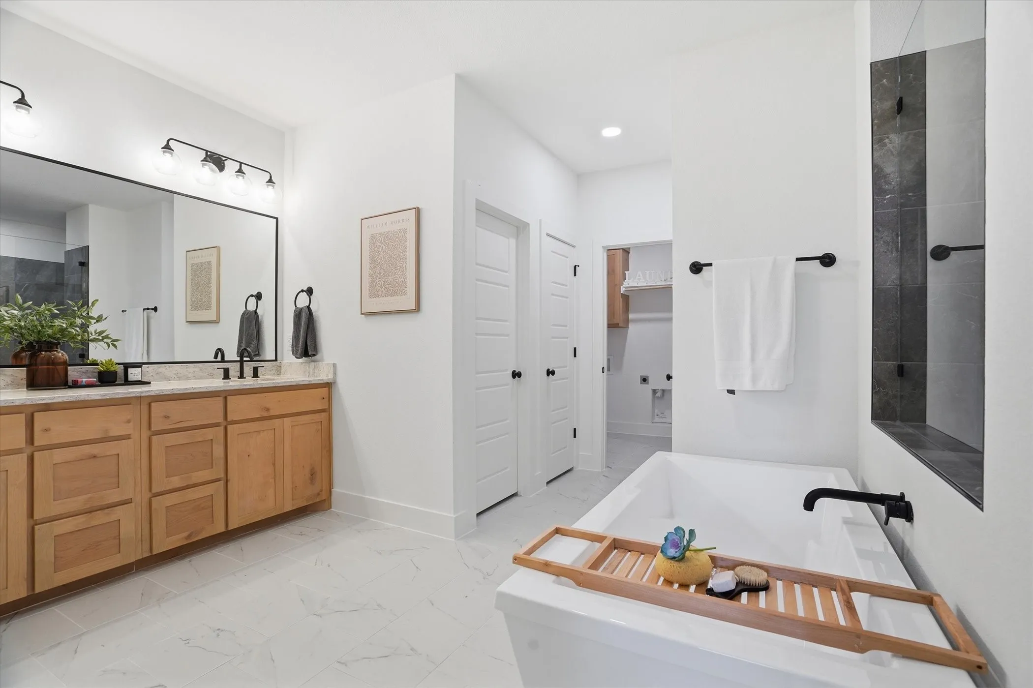 Freestanding soaking tub with dark-finish fixtures, dual vanity with wood cabinetry, large mirror, and white tile flooring