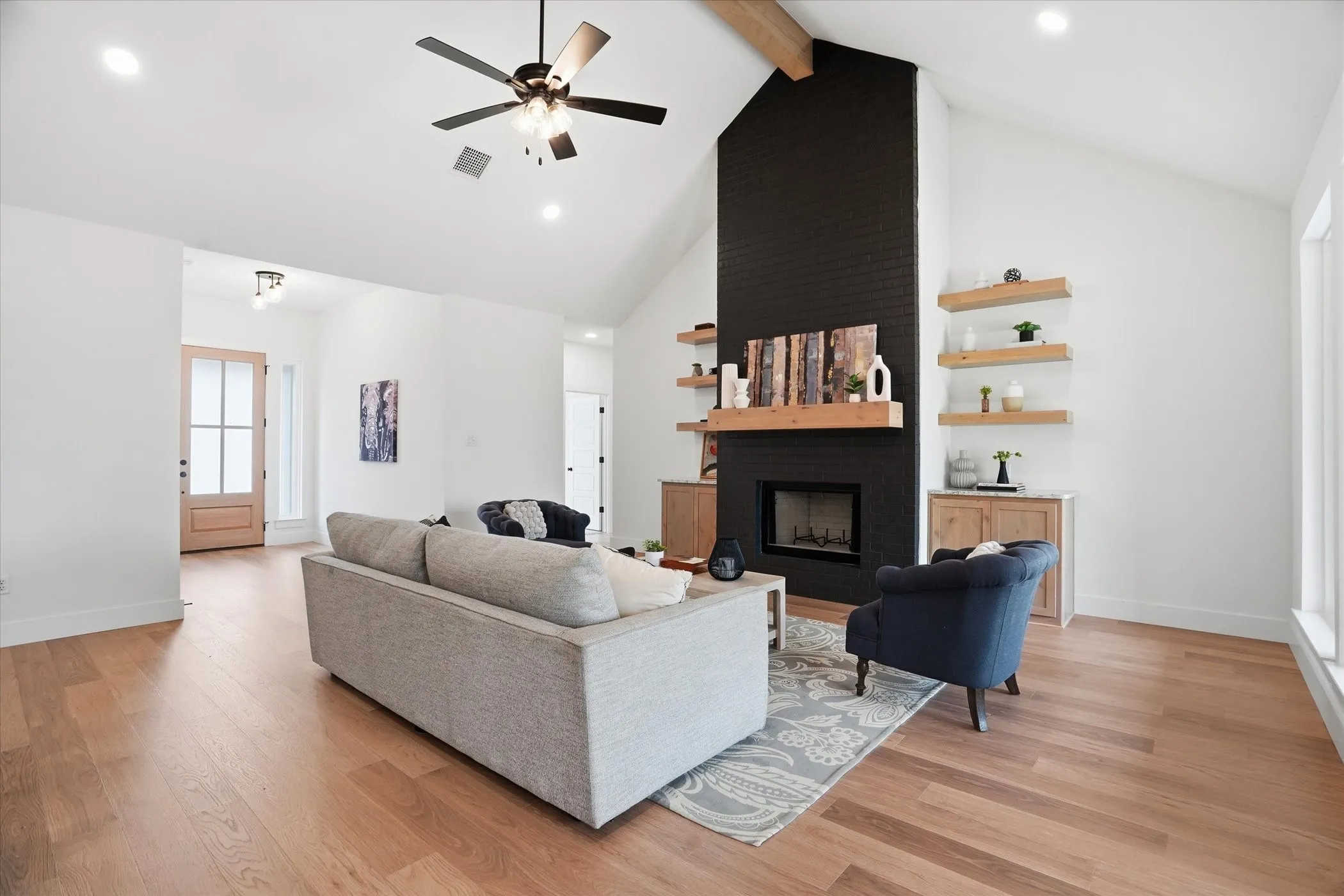 Vaulted ceiling with exposed beam, wood-finish flooring, black brick fireplace surround, wood mantel, and integrated wood shelving