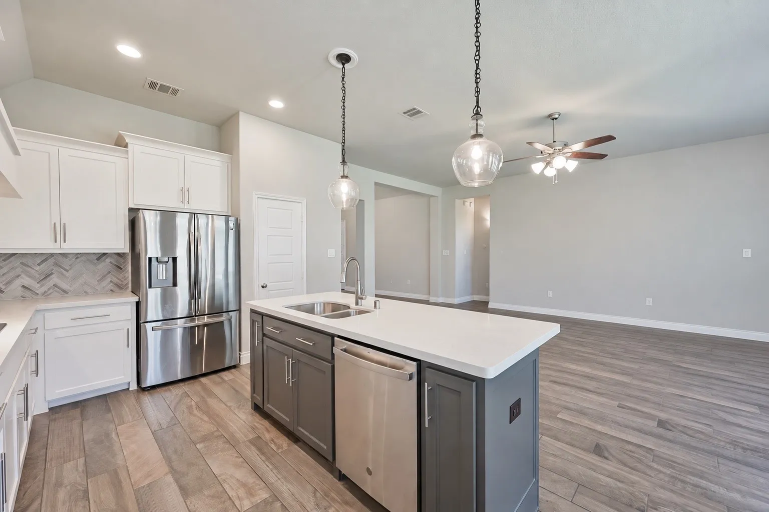 Kitchen island with a built-in stainless steel sink, sleek stainless steel appliances, white cabinetry with silver hardware, gray subway tile backsplash, and wood-finish flooring