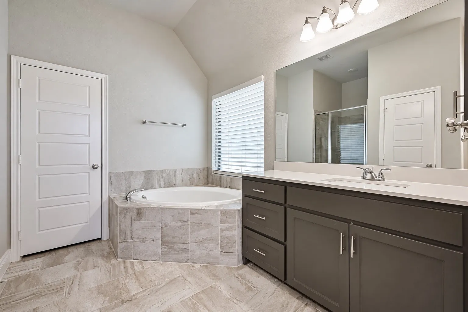 Spacious bathroom featuring a built-in soaking tub with tiled surround, a large vanity with ample storage, a wide mirror, and a modern light fixture