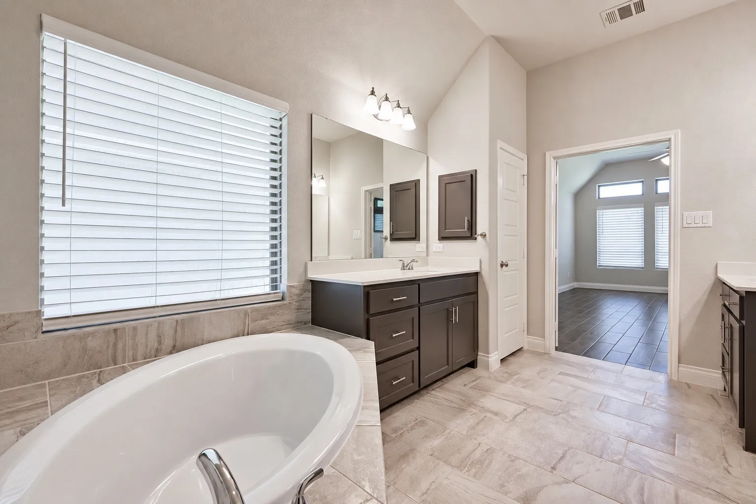 Freestanding soaking tub, dual vanities with white countertops, tiled flooring, and a window with blinds