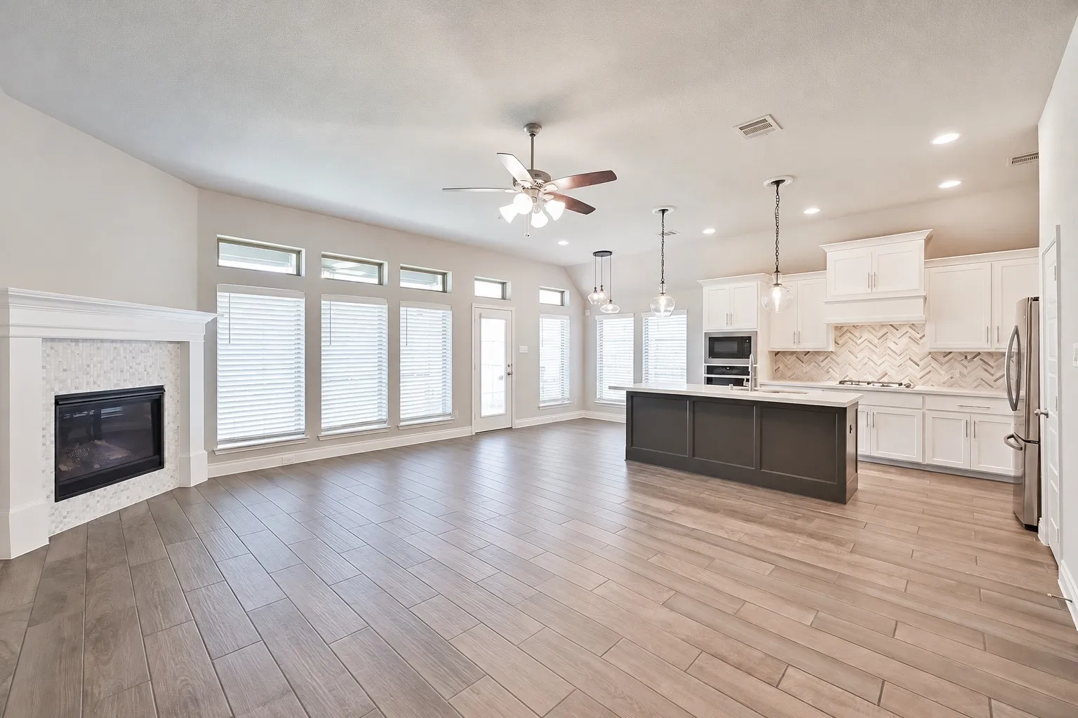 Spacious open-concept layout featuring wood-finish flooring, a built-in fireplace with a tiled surround, a kitchen island with pendant lighting, white cabinetry, and stainless steel appliances