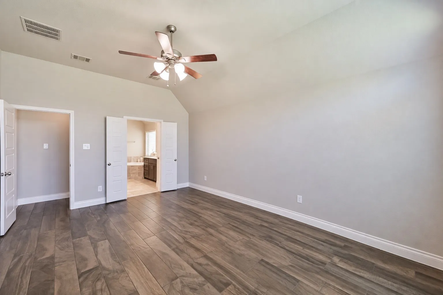 Vaulted ceiling with a ceiling fan, wood-finish flooring, light gray wall tones, and double doors opening to a full bathroom featuring a built-in tub