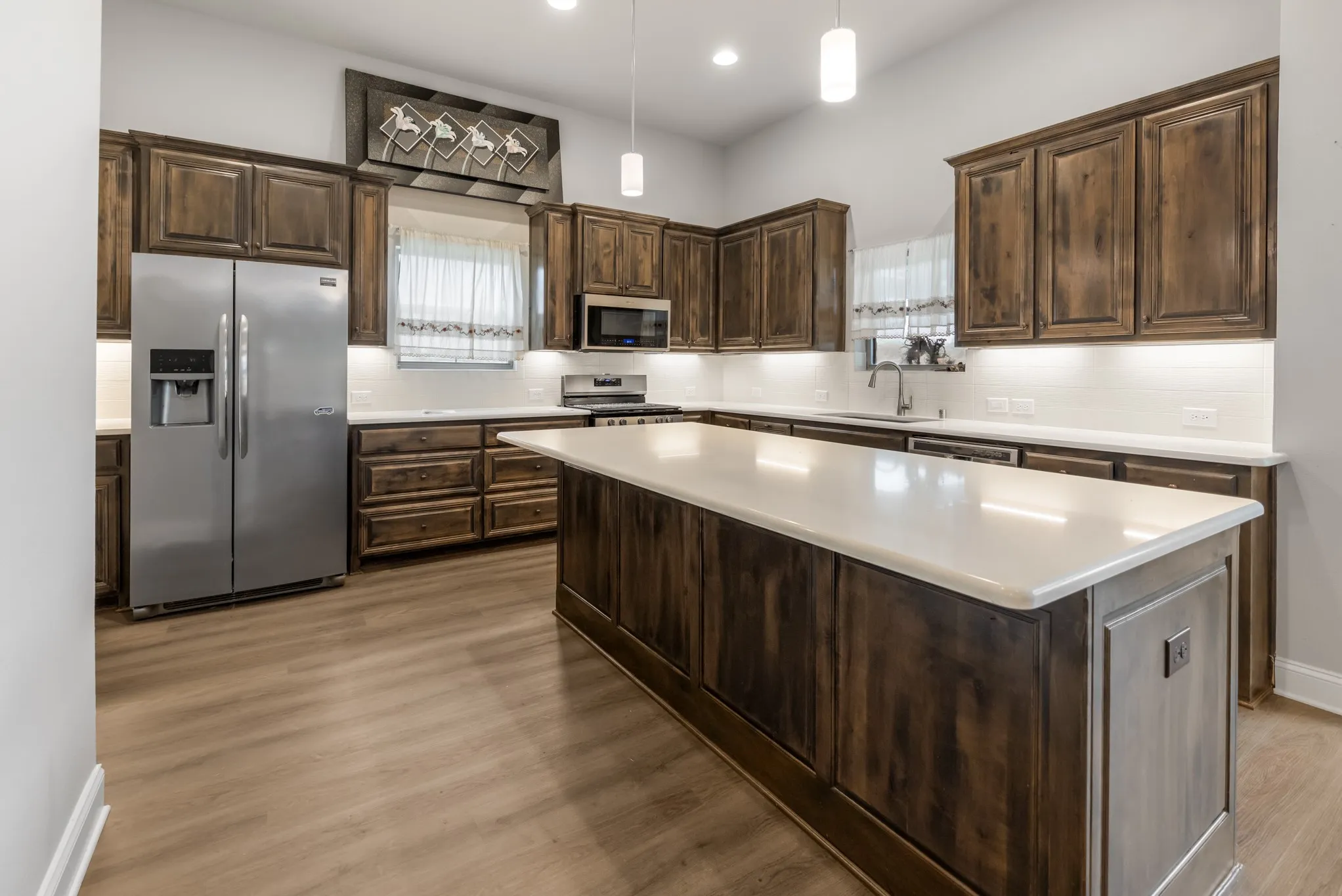 Spacious kitchen featuring wood-finish flooring, dark wood cabinetry, a large center island with a light-toned countertop, stainless steel appliances, and a subway tile backsplash