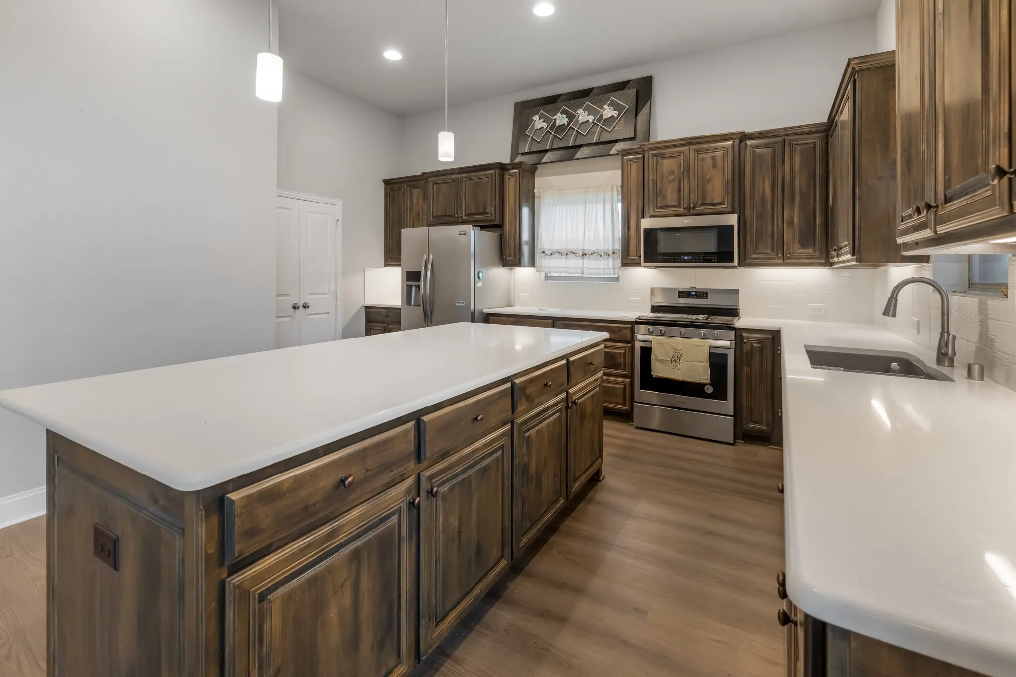 Expansive kitchen island with a light-toned countertop, wood-finish cabinetry, stainless steel appliances, and wood-finish flooring