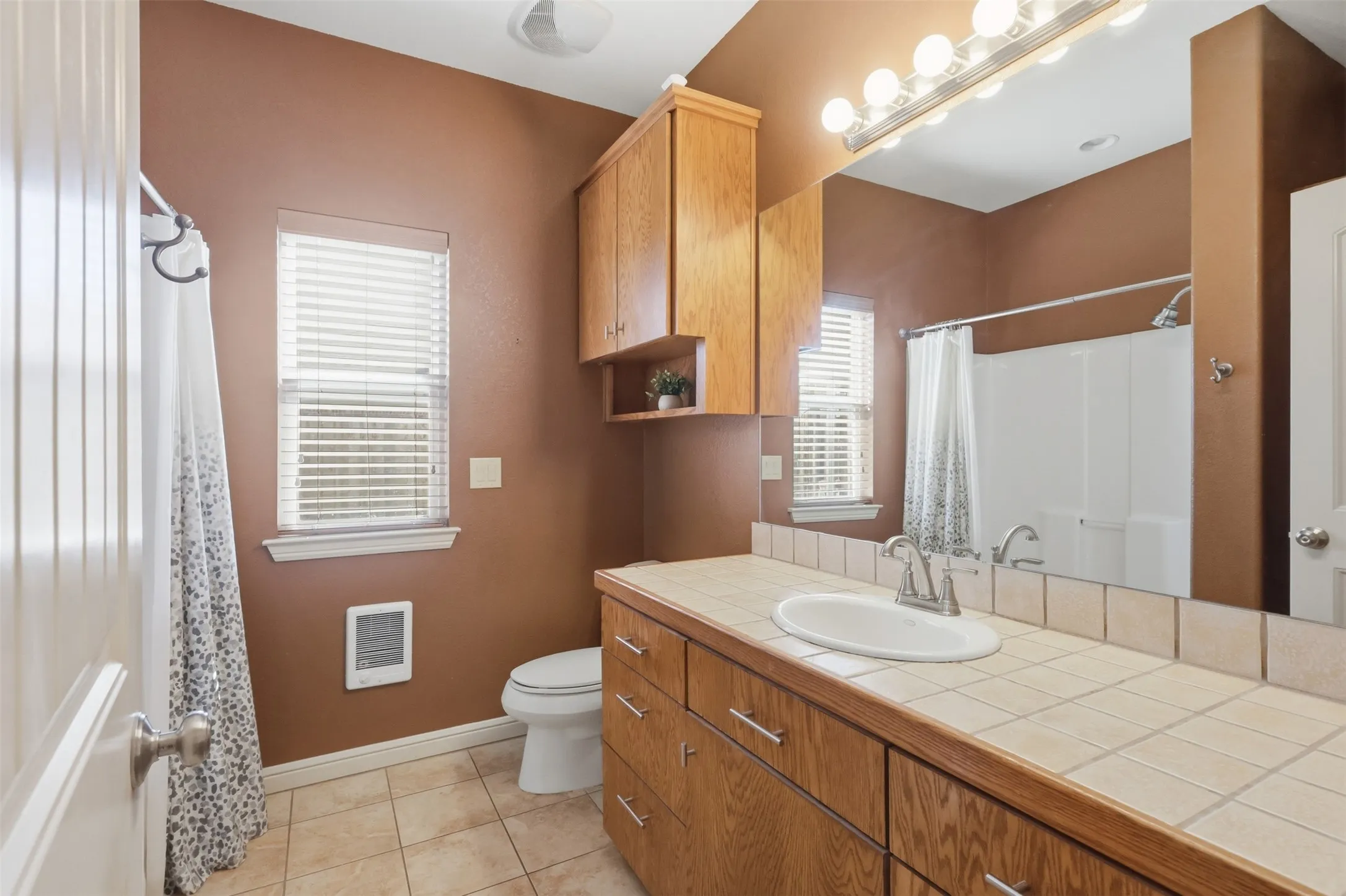 Warm-toned bathroom featuring a tile-top vanity with integrated sink, wood-finish cabinetry, and a full-width mirror