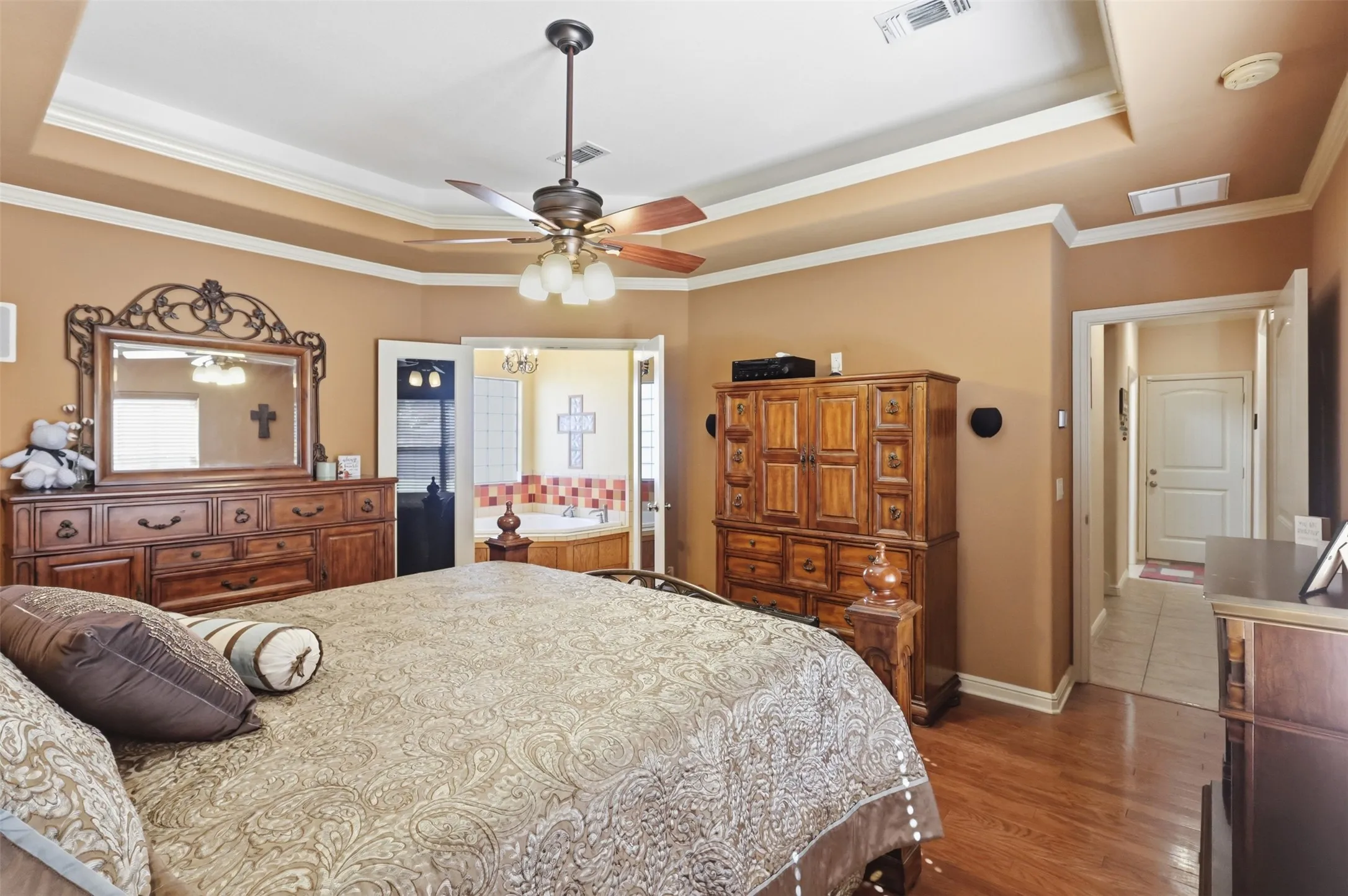 Recessed tray ceiling with crown molding, wood-finish flooring, ceiling fan with integrated lighting, and an alcove with a built-in jetted tub