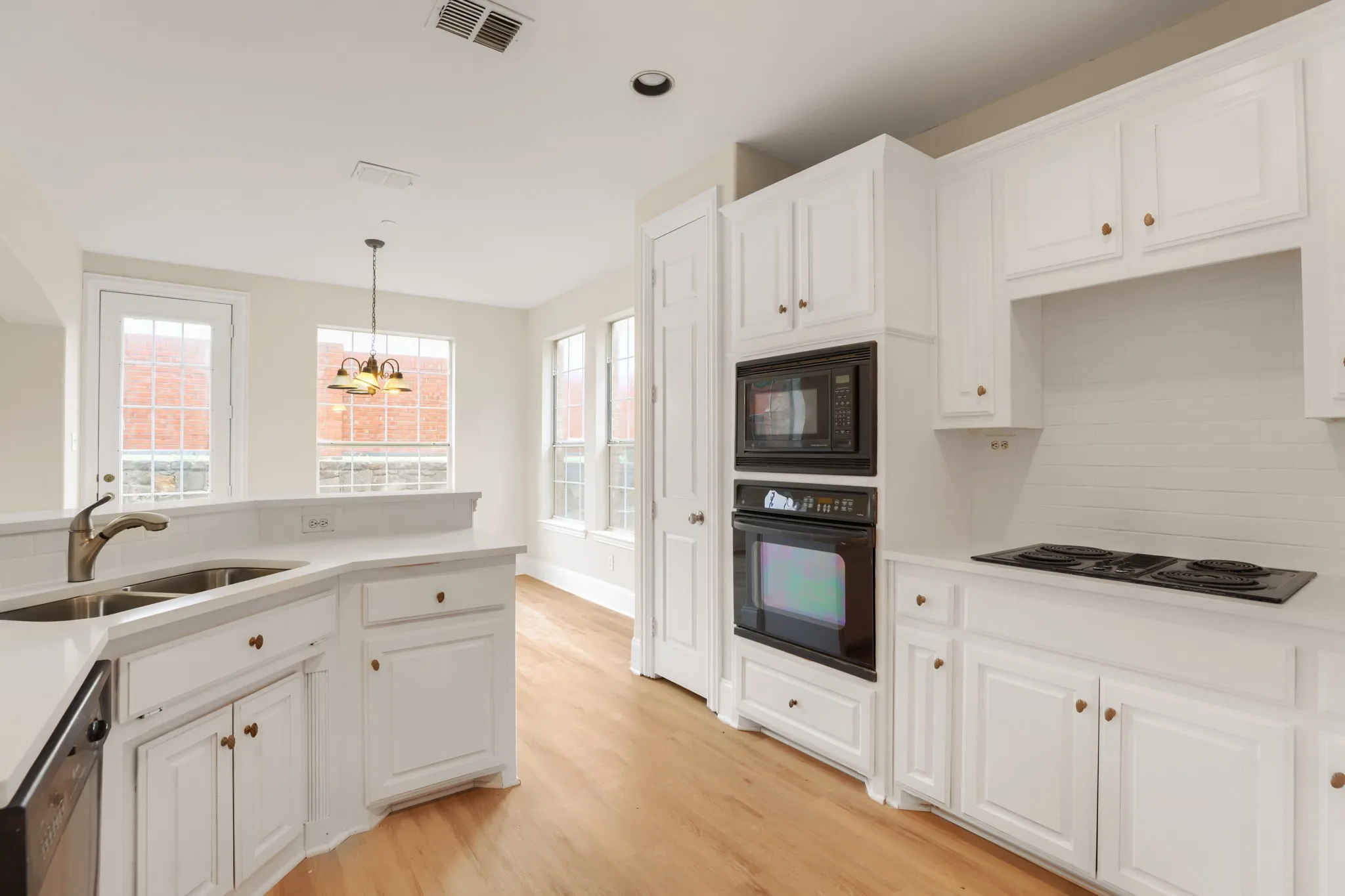 Kitchen featuring light wood-finish flooring, white cabinetry with brass-toned hardware, double basin sink with a gooseneck faucet, built-in microwave, and an electric cooktop
