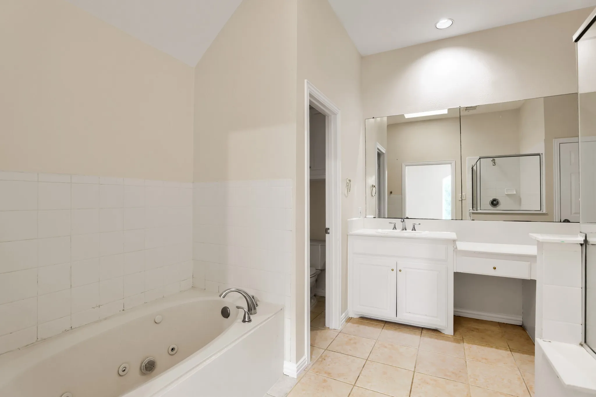 Jetted soaking tub, white tile wainscoting, white vanity with integrated sink, extensive wall-mounted mirror, and tiled flooring