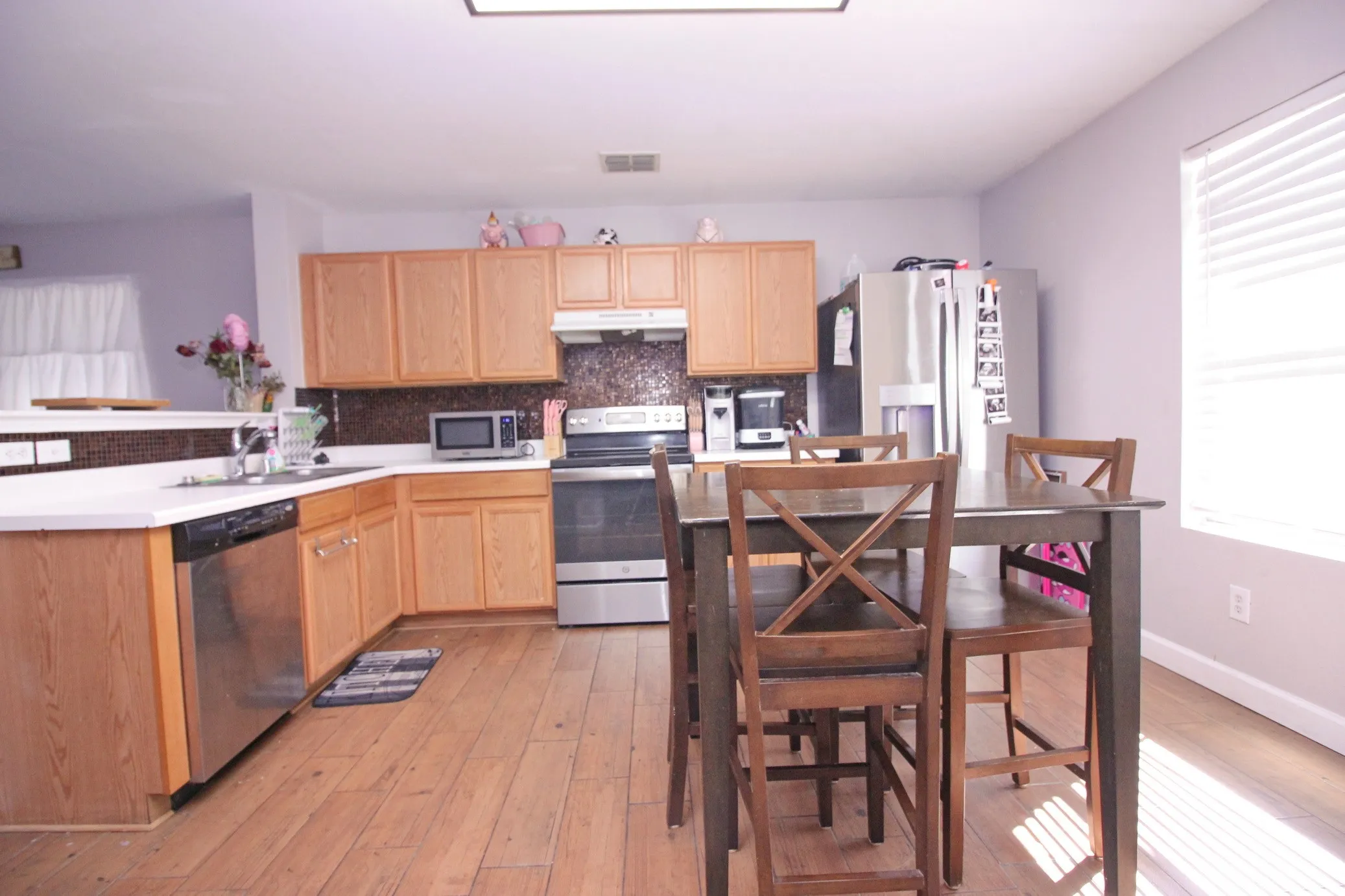 Wood-finish flooring, light wood cabinetry, white countertops, dark mosaic tile backsplash, and stainless steel appliances
