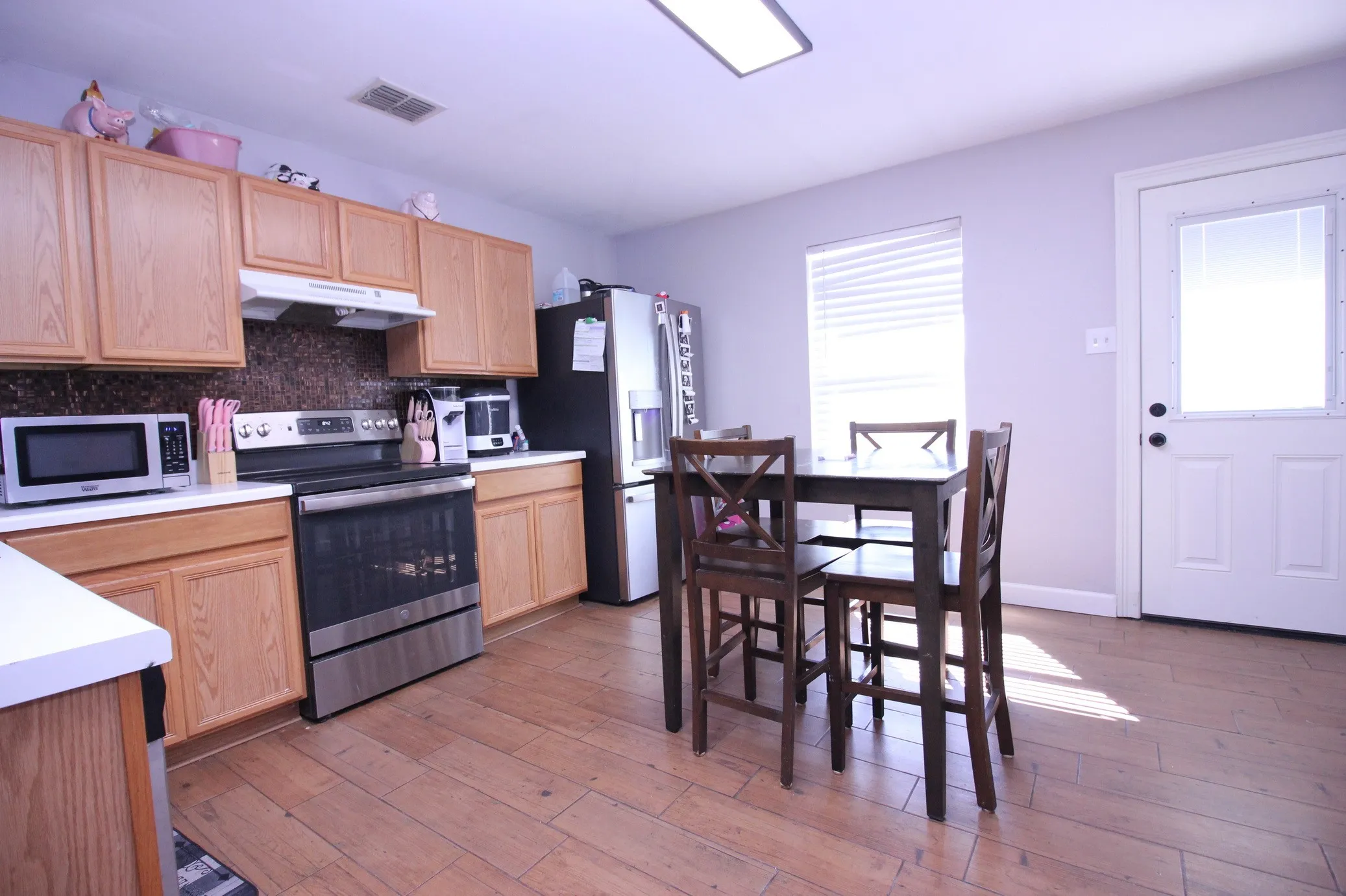 Wood-finish flooring, light wood cabinetry, white countertops, dark mosaic tile backsplash, and a window with blinds
