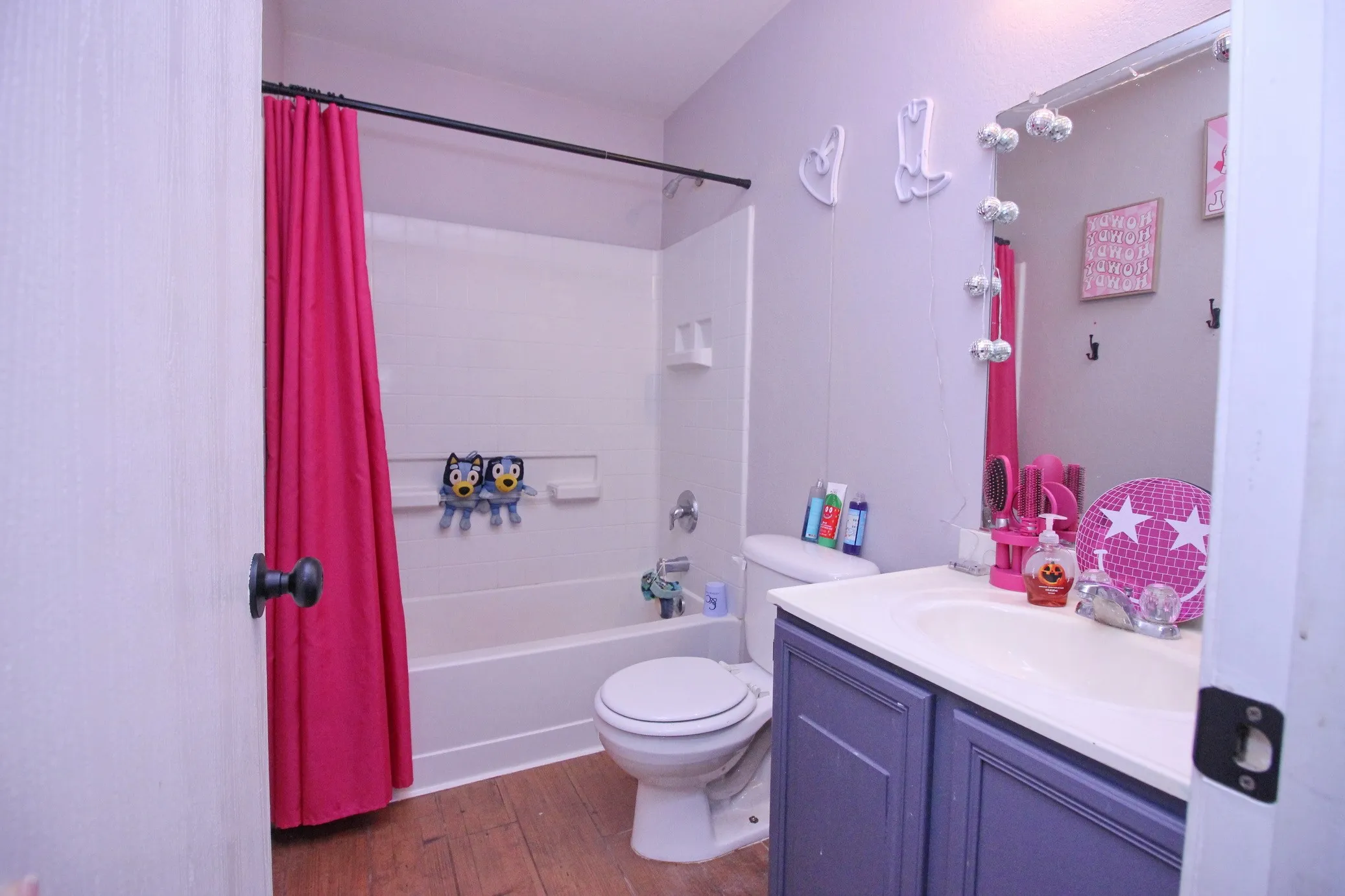 Bathroom featuring wood-finish flooring, a white bathtub with shower, a toilet, a vanity with a white countertop and integrated sink, and a framed mirror