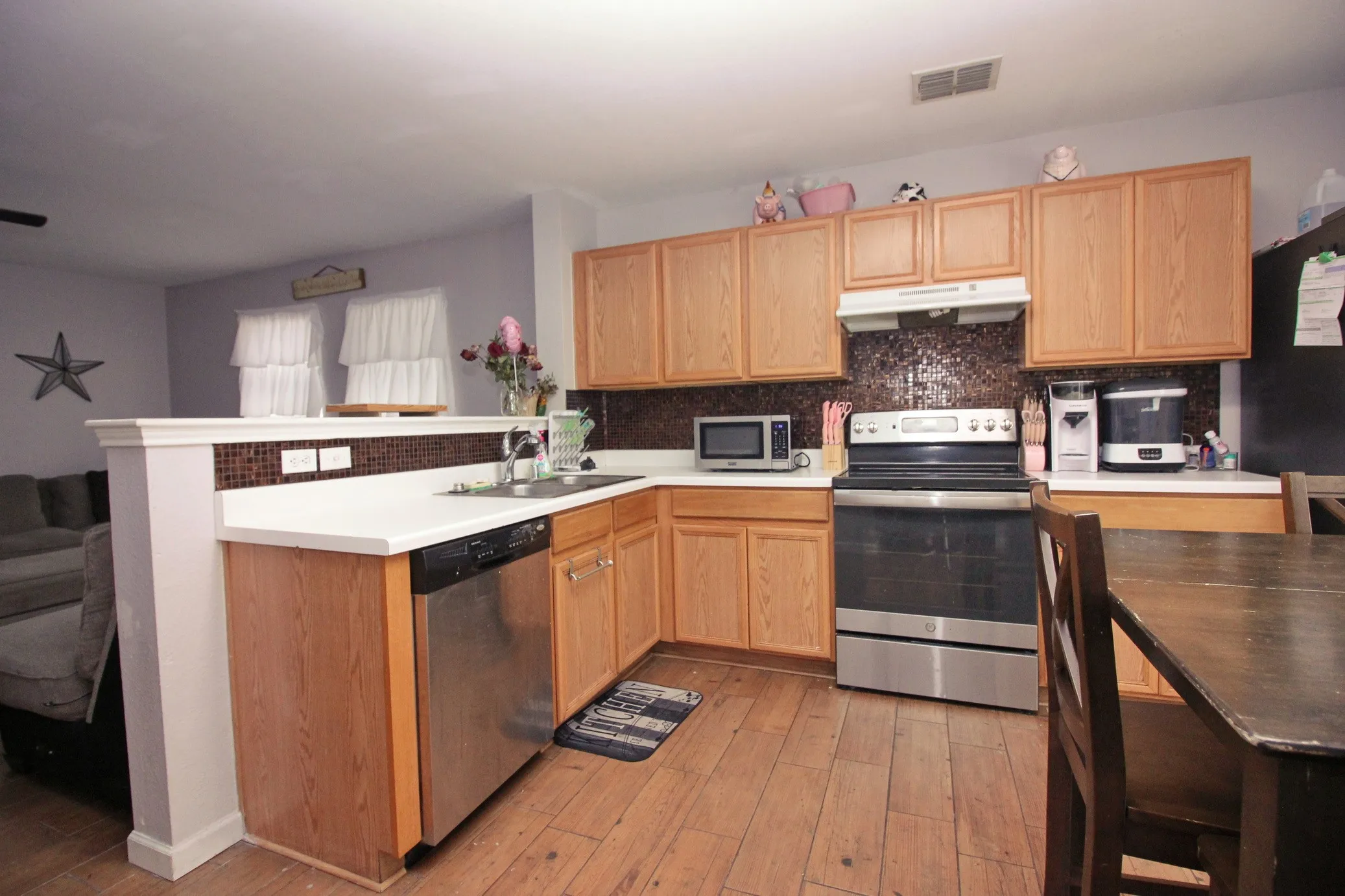 Contemporary kitchen featuring wood-finish flooring, stainless steel appliances, light wood-finish cabinetry, white countertops, and a mosaic tile backsplash