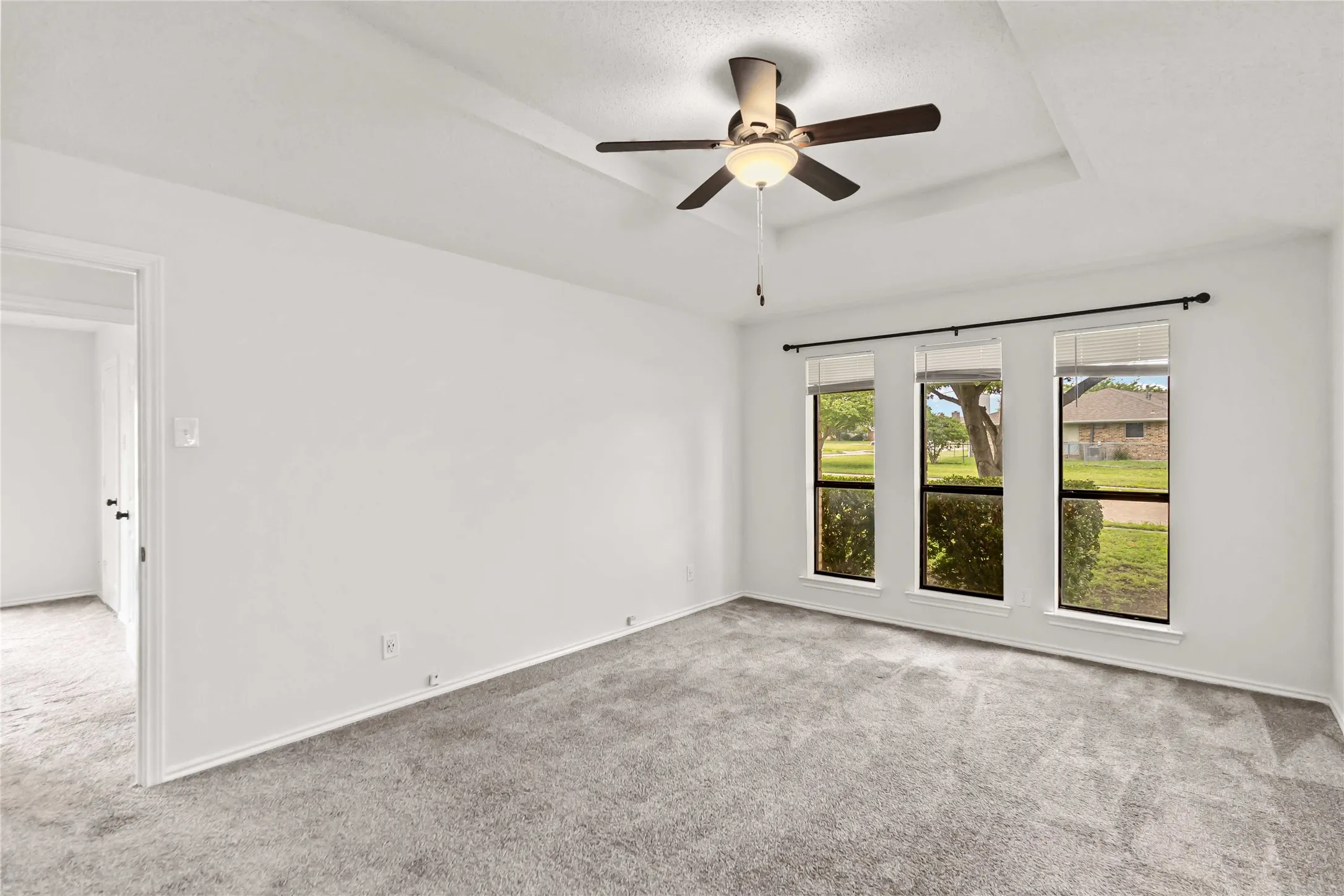 Room featuring light gray carpet, three windows with dark frames, a tray ceiling, a ceiling fan with light, and white walls