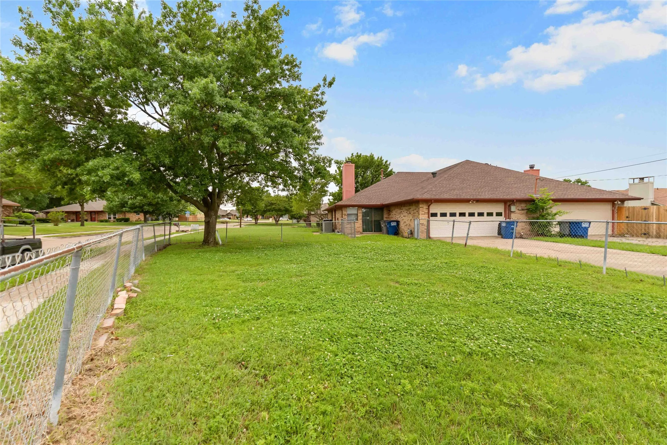 Expansive lawn area featuring a mature tree, chain-link fencing, and a paved driveway offering access to the attached two-car garage