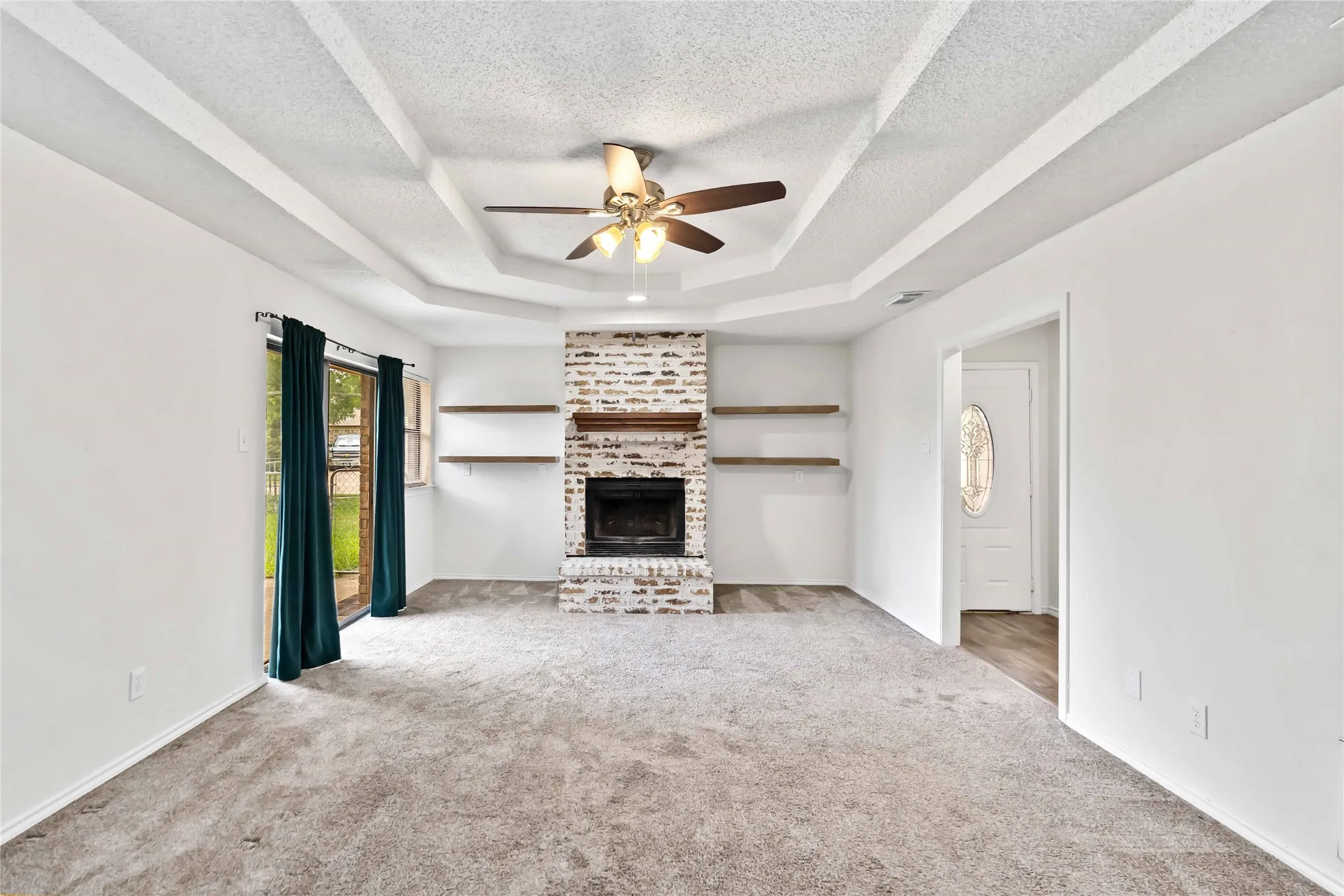 Living area featuring a white brick fireplace with wood mantel and open shelving, gray carpet, a tray ceiling with a ceiling fan, and a sliding glass door
