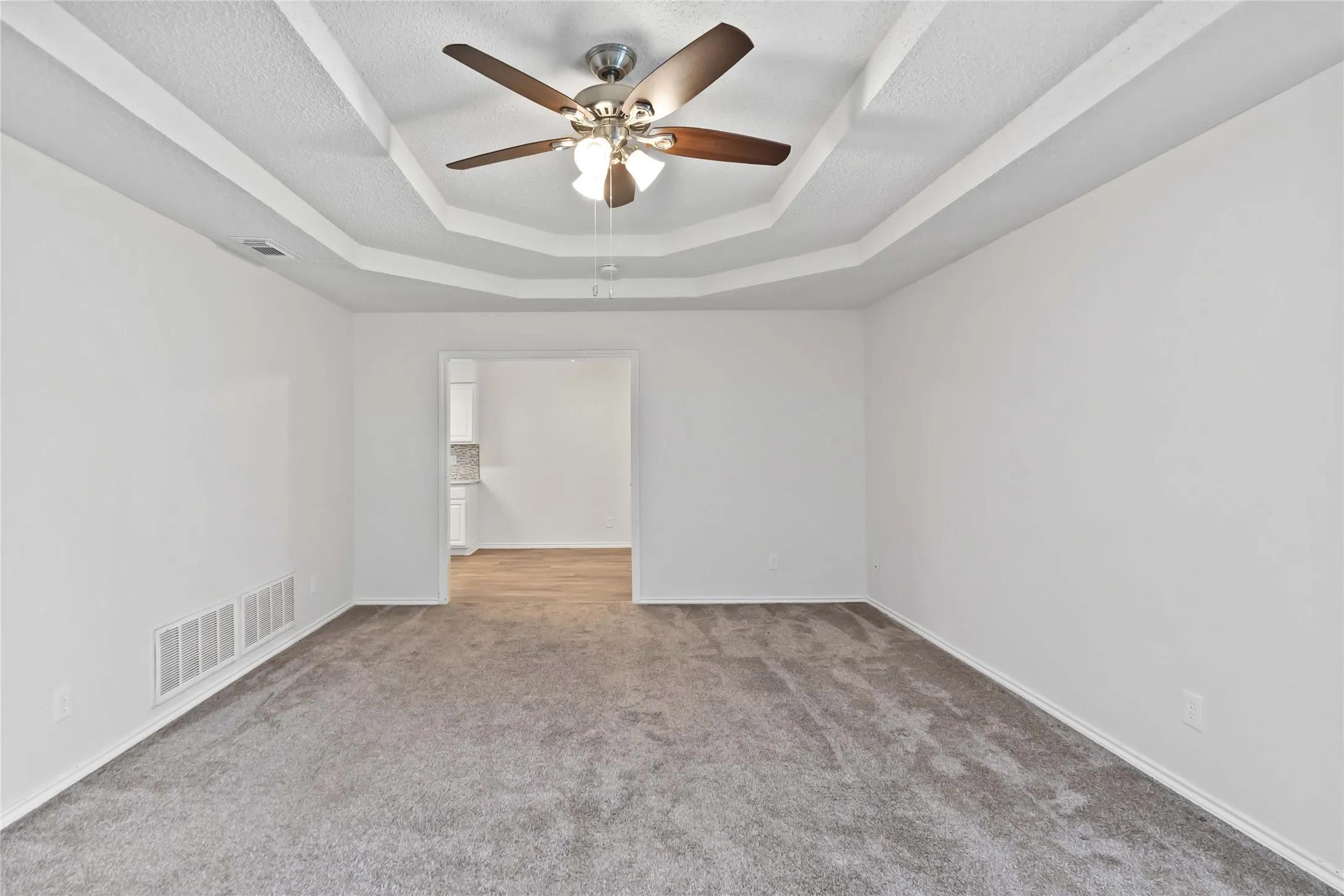 Recessed tray ceiling with a ceiling fan, plush gray carpeting, and white baseboards
