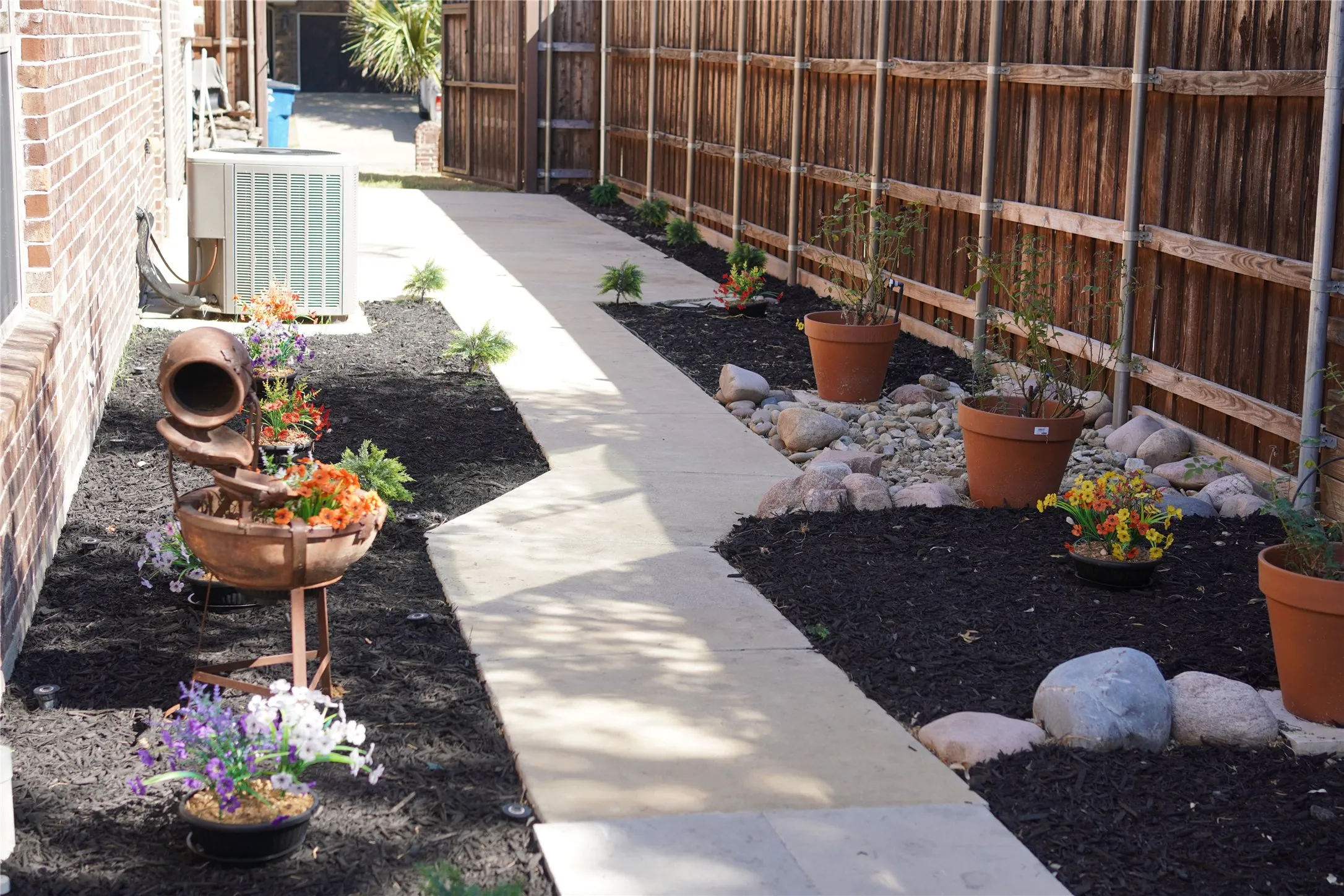 Side yard featuring a concrete pathway, a wooden privacy fence, dark mulch beds, and decorative rocks