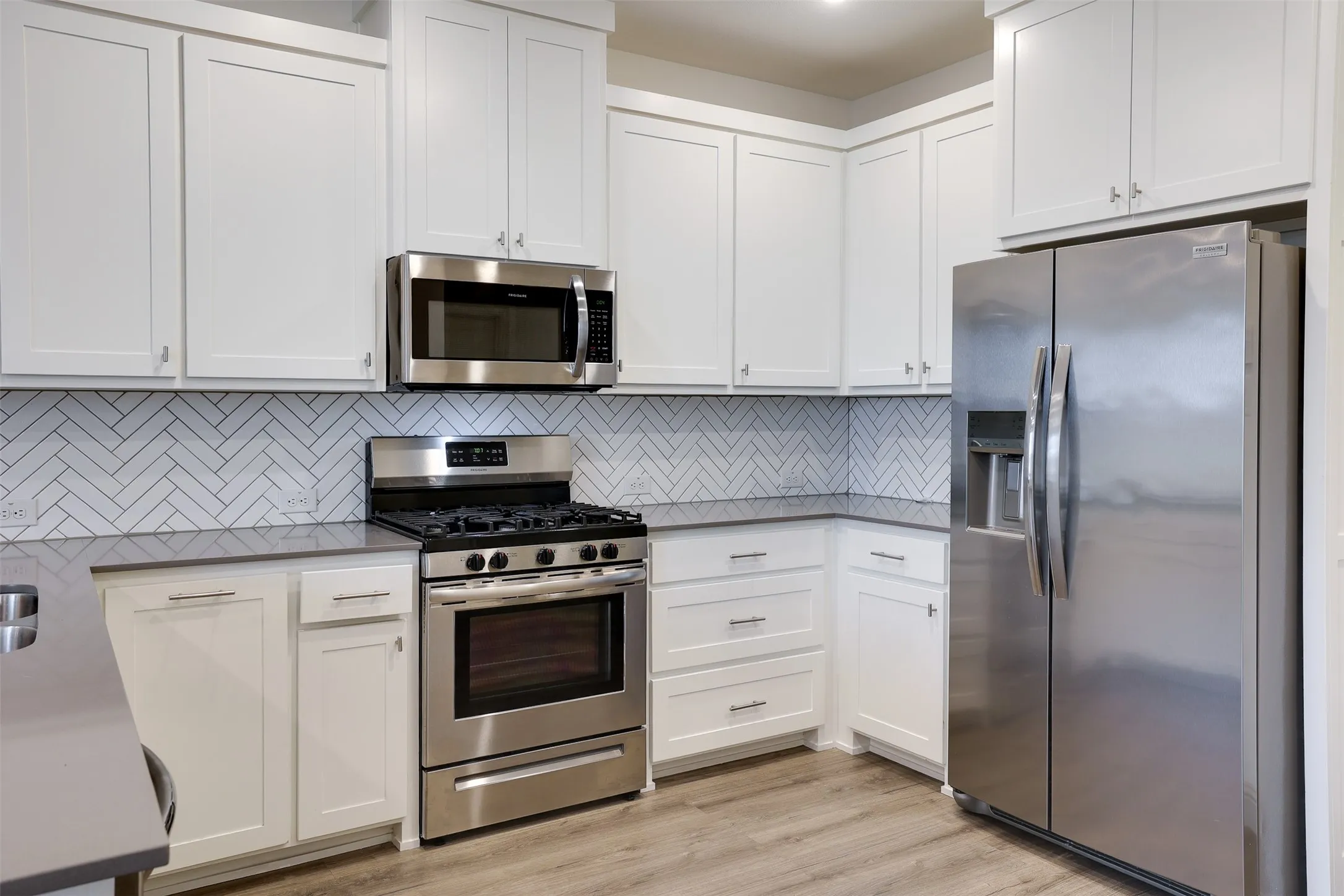 White shaker cabinetry, herringbone tile backsplash, stainless steel appliances, dark countertops, and wood-finish flooring