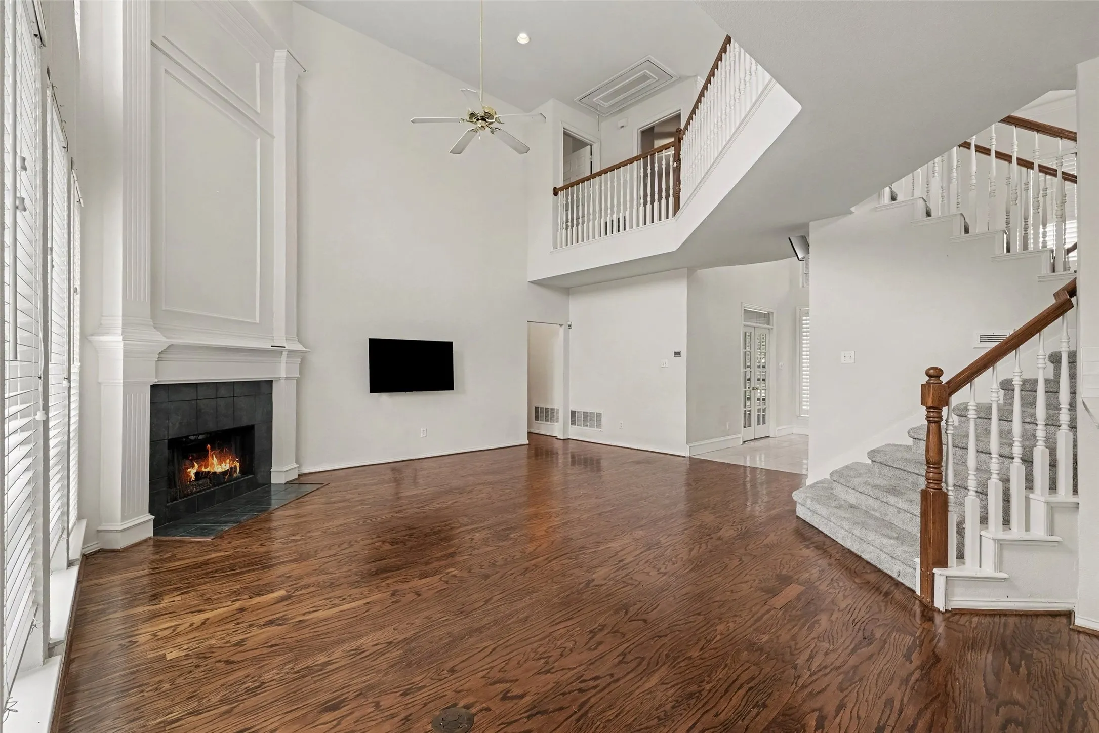 Hardwood flooring throughout, a soaring two-story ceiling, a fireplace with a stone surround, and an open staircase with a carpeted runner