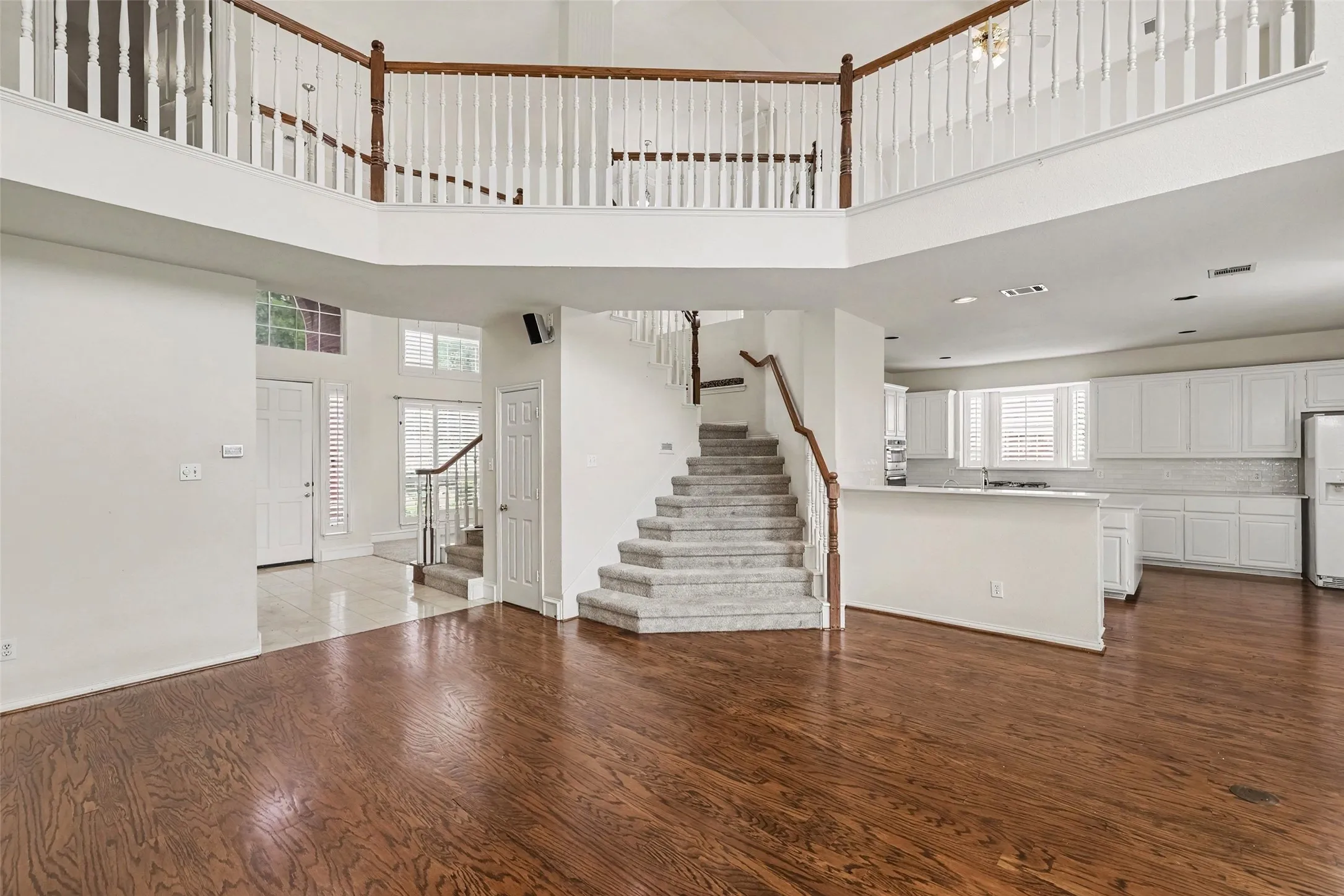 Hardwood flooring, two-story entryway, grand staircase with wood railing, white kitchen cabinetry, and a second-story interior balcony