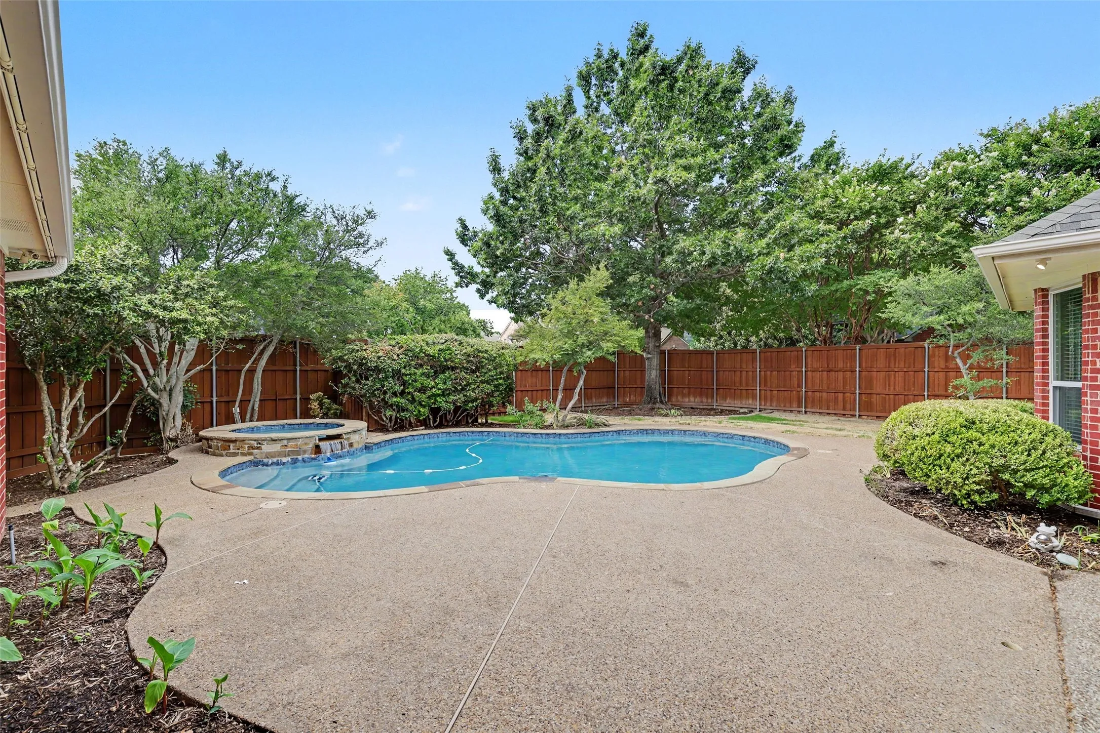 Backyard featuring a free-form swimming pool with integrated spa, concrete decking, wood fencing, and mature landscaping