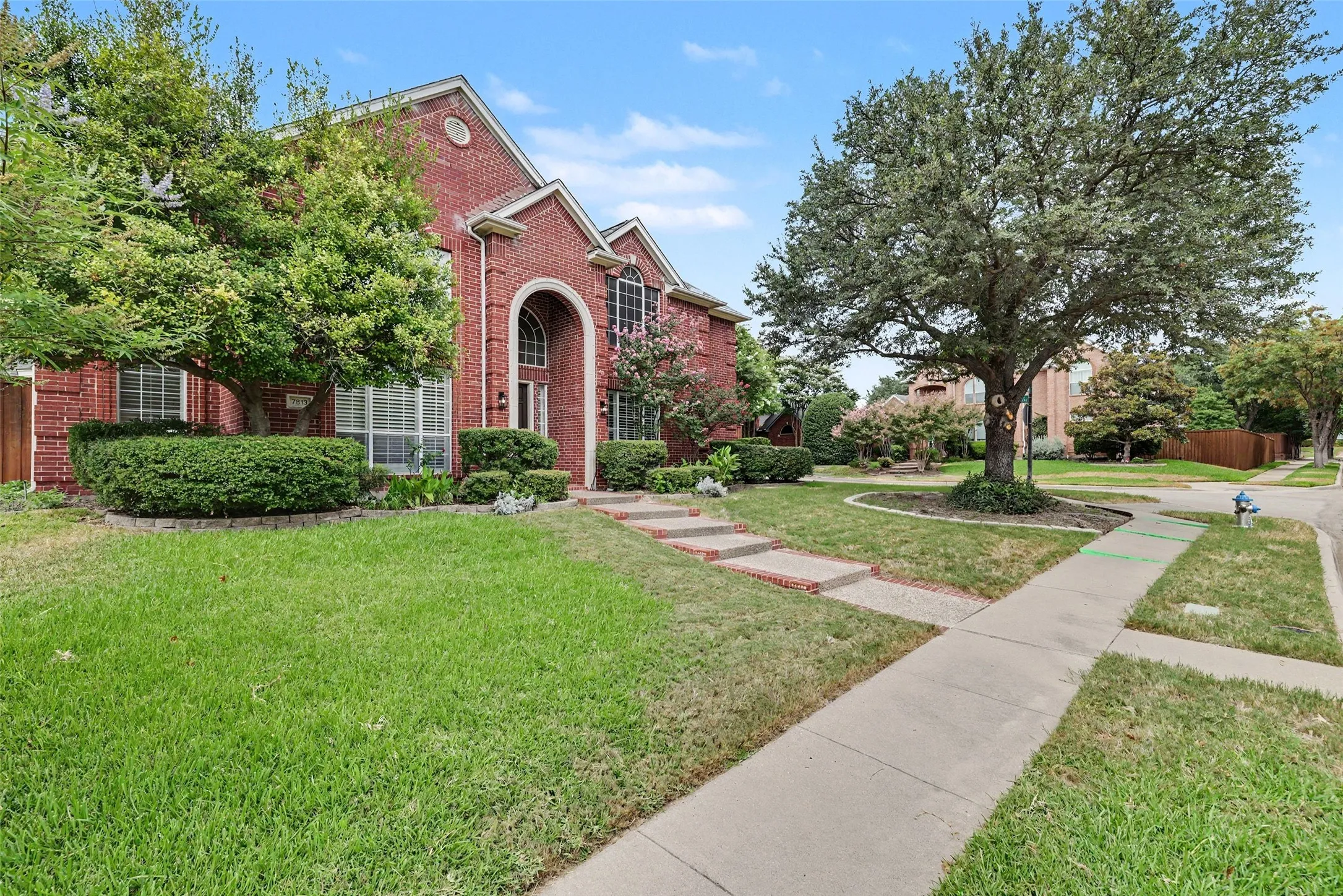 Red brick exterior with an arched entry, mature trees, landscaped shrubbery, and a concrete sidewalk