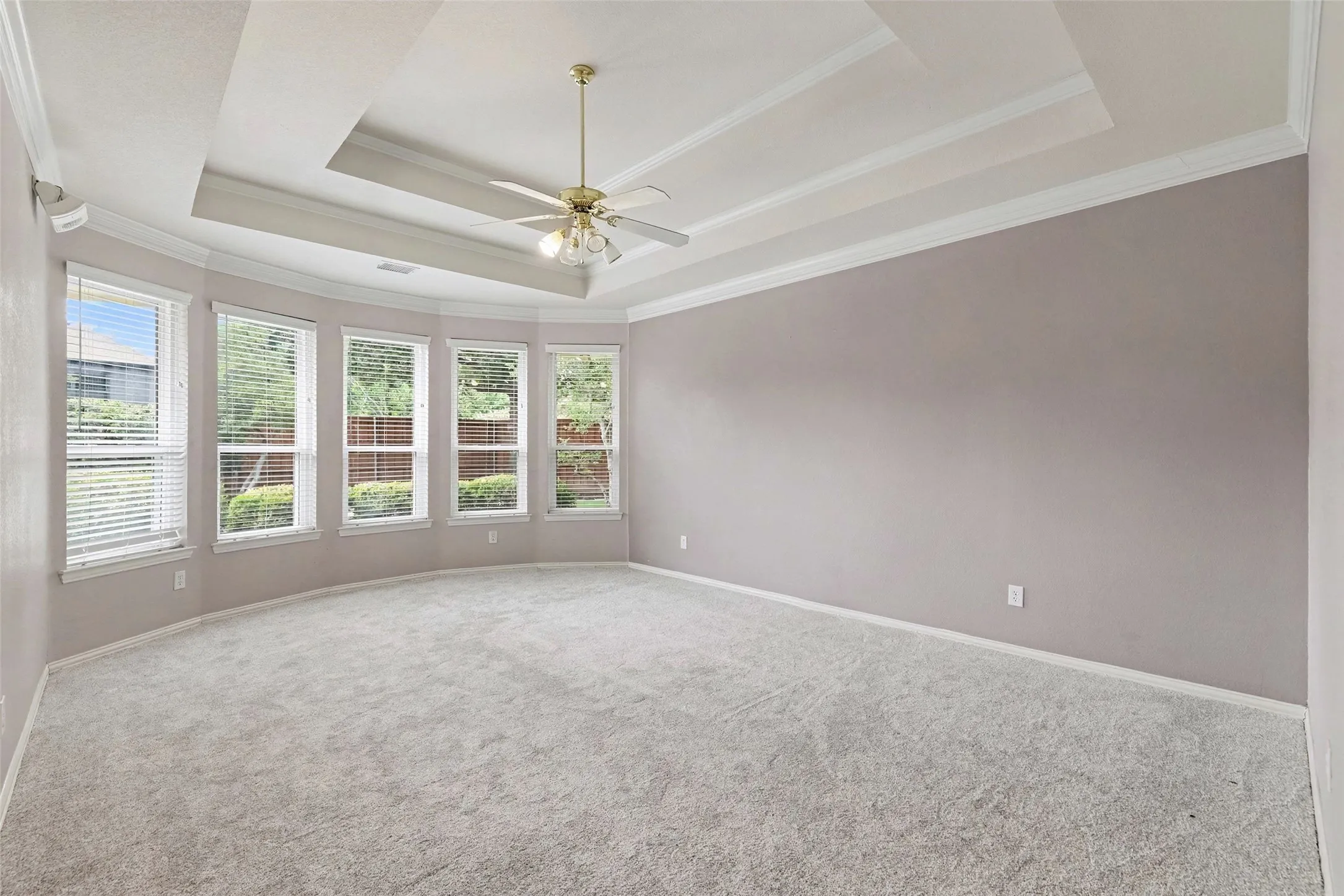 Recessed tray ceiling with crown molding, ceiling fan with integrated lighting, multiple windows with blinds, and light-toned carpet flooring