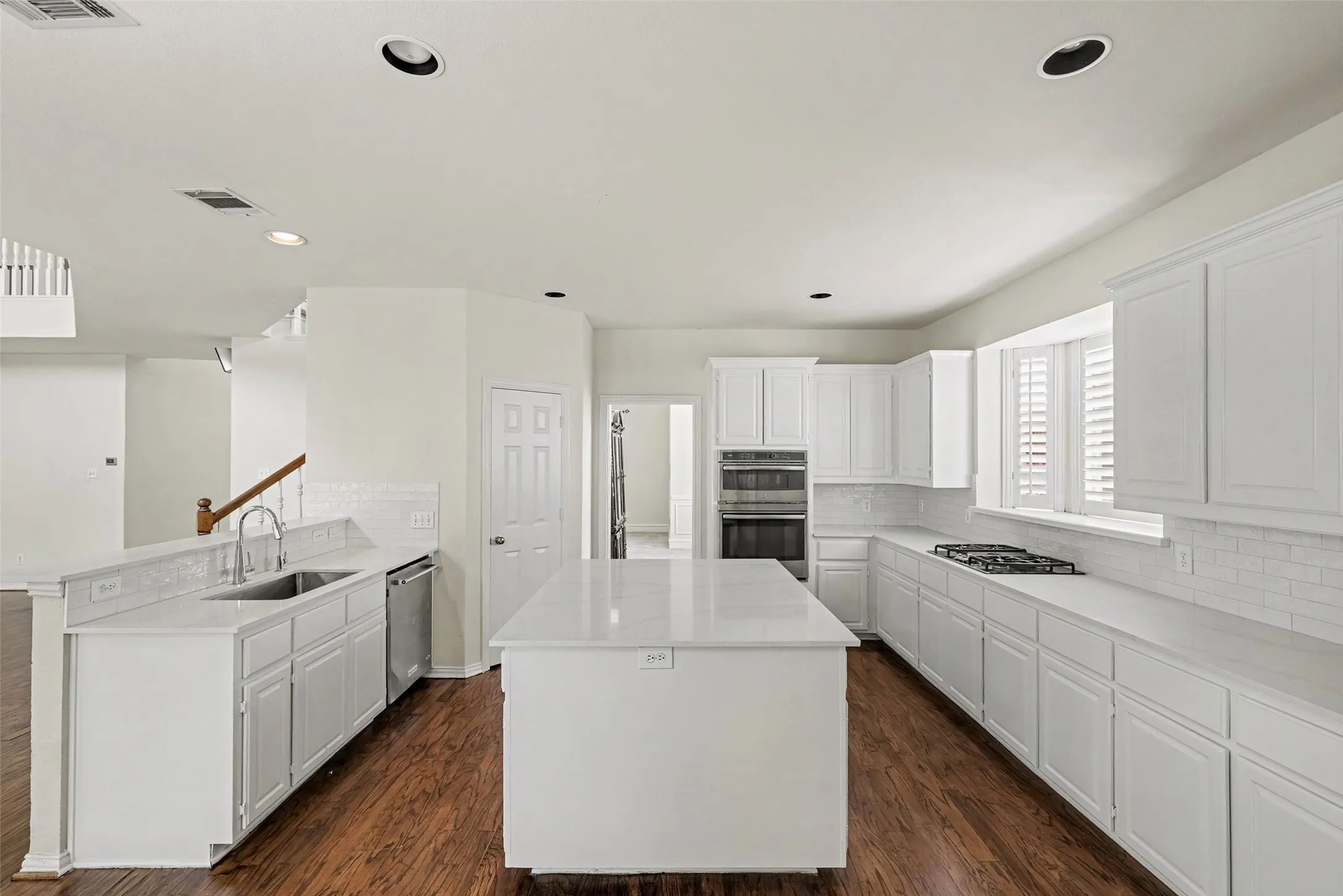 Chef's kitchen featuring white cabinetry, a large center island with white quartz countertop, stainless steel appliances including dual wall ovens and gas cooktop, white subway tile backsplash, and wood-finish flooring throughout