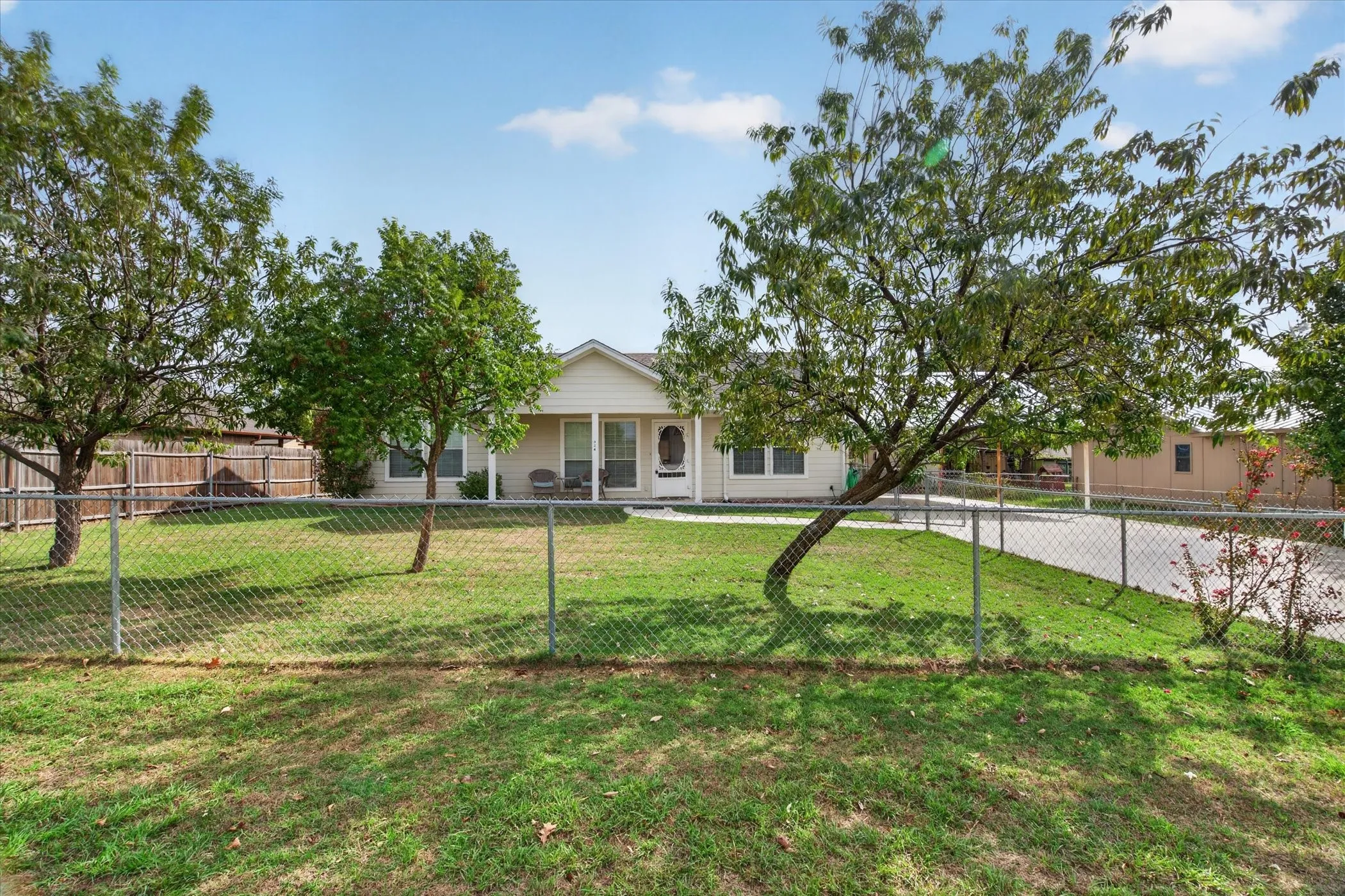 Single-story exterior featuring light-toned horizontal siding, a covered front porch, mature trees, and a chain-link fenced perimeter