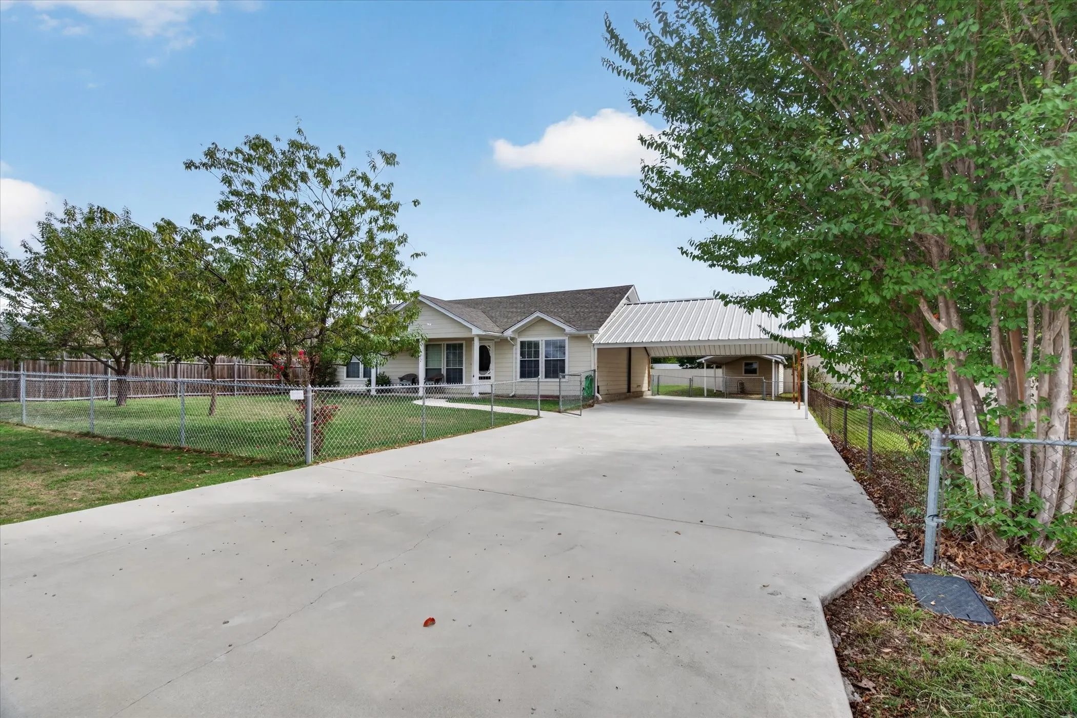 Single-story exterior featuring light-colored siding, a covered carport with metal roofing, an expansive concrete driveway, and a chain-link fenced yard
