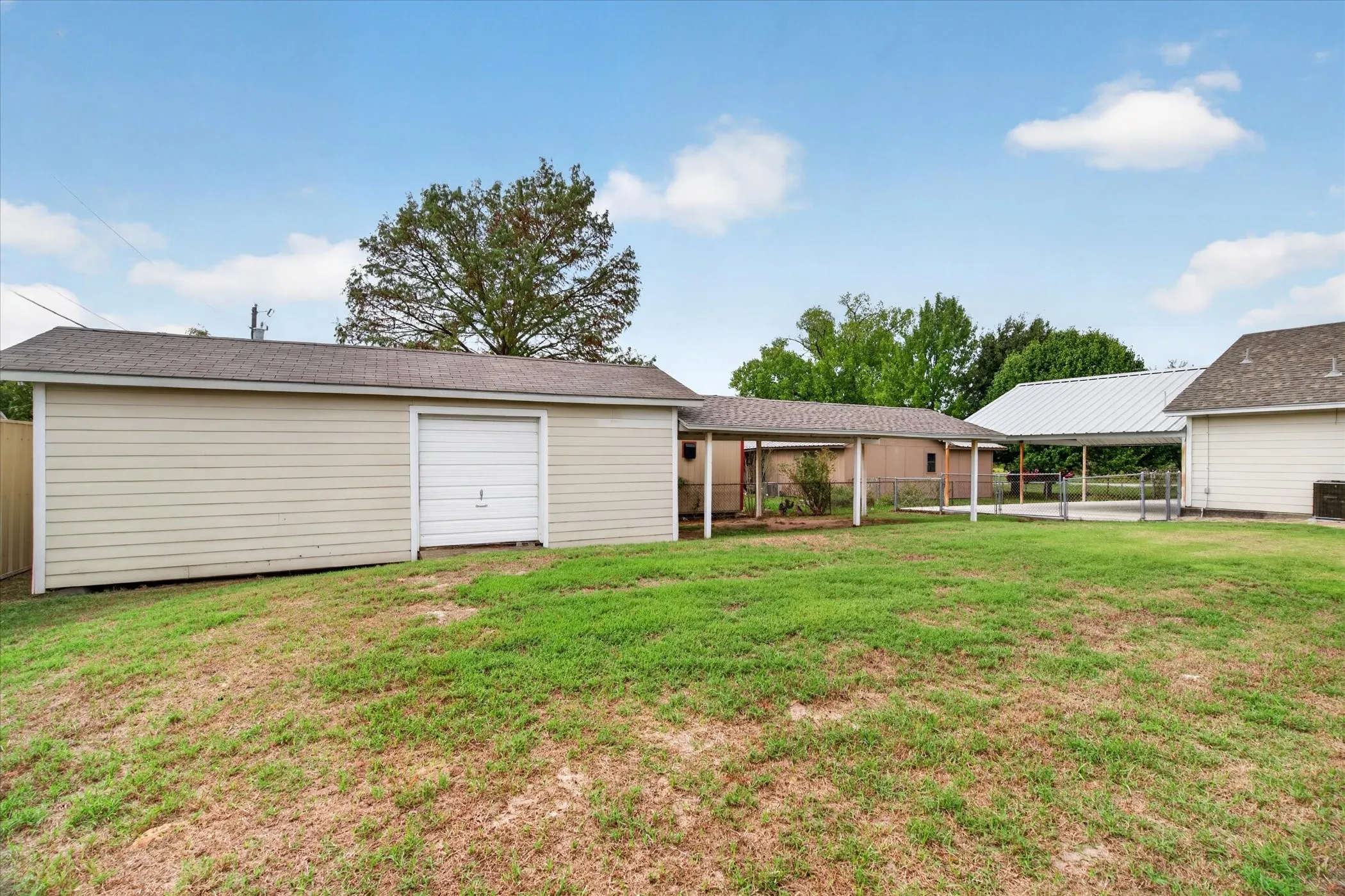 Detached garage with horizontal siding and shingled roof, accompanied by a covered carport and expansive lawn area