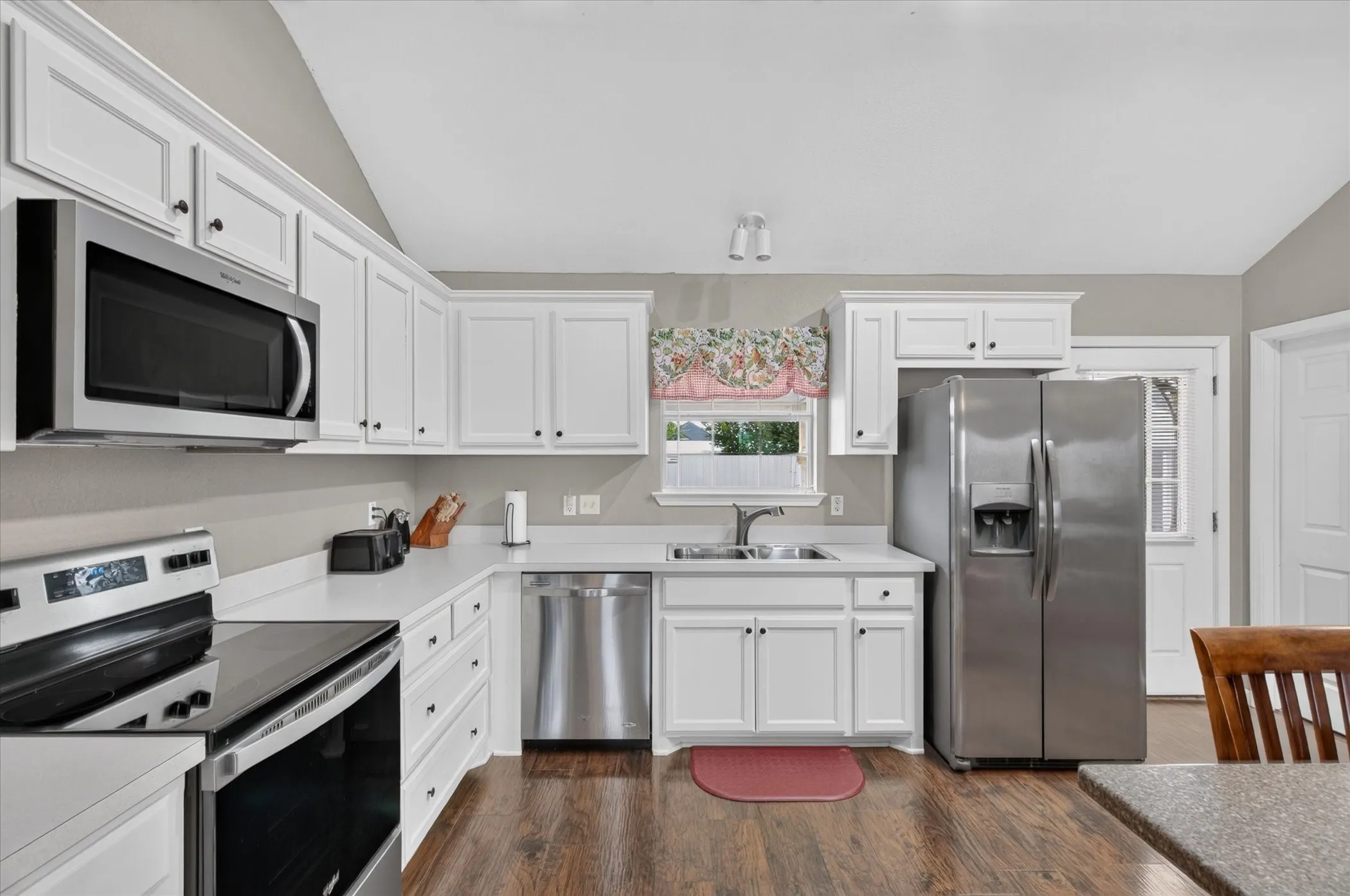 Kitchen featuring white shaker-style cabinetry, stainless steel appliances, wood-finish flooring, solid-surface countertops, and a vaulted ceiling