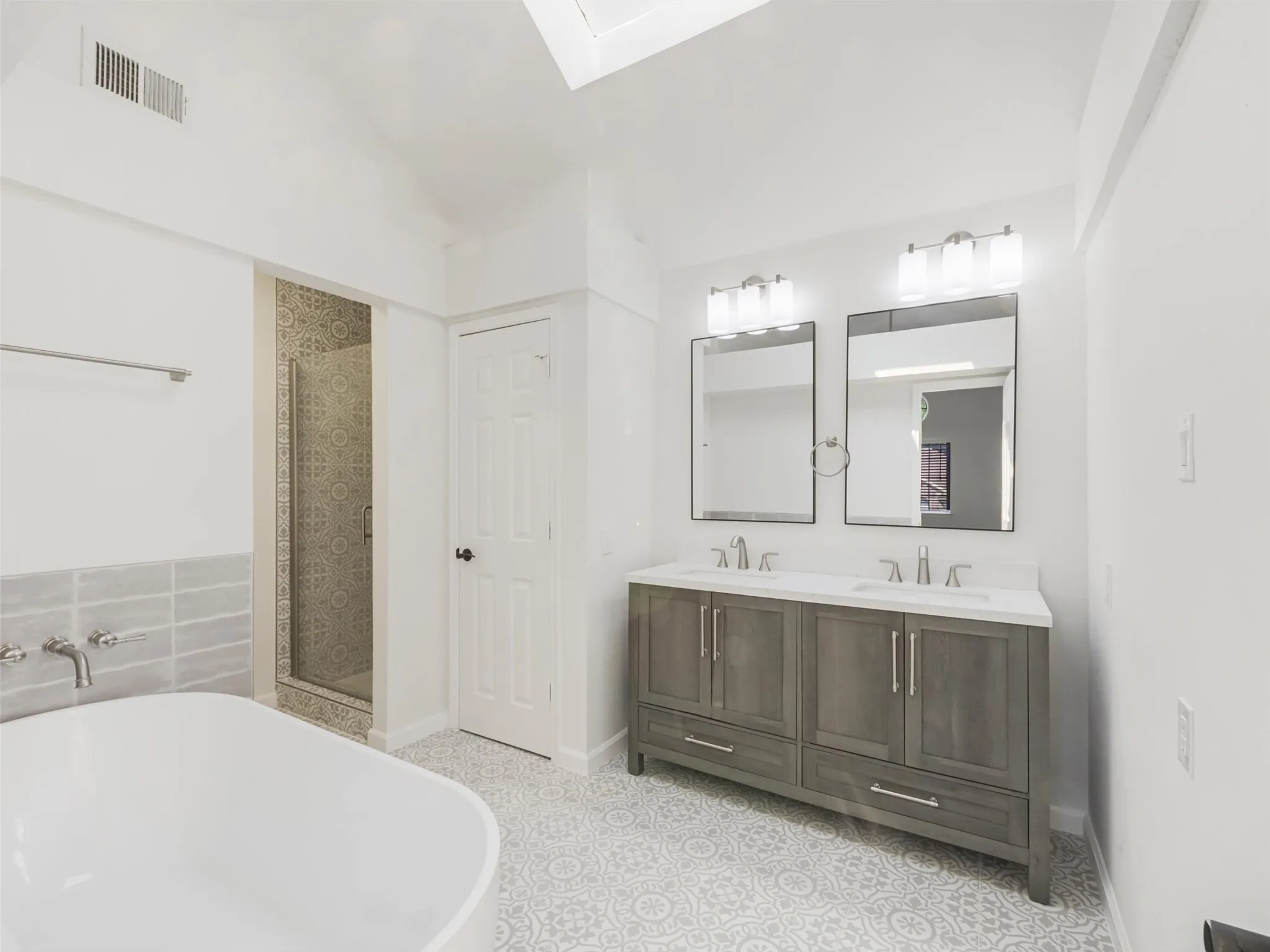 Spacious bathroom featuring a double vanity with ample storage, a freestanding soaking tub, a glass-enclosed shower, patterned floor tiling, and a skylight
