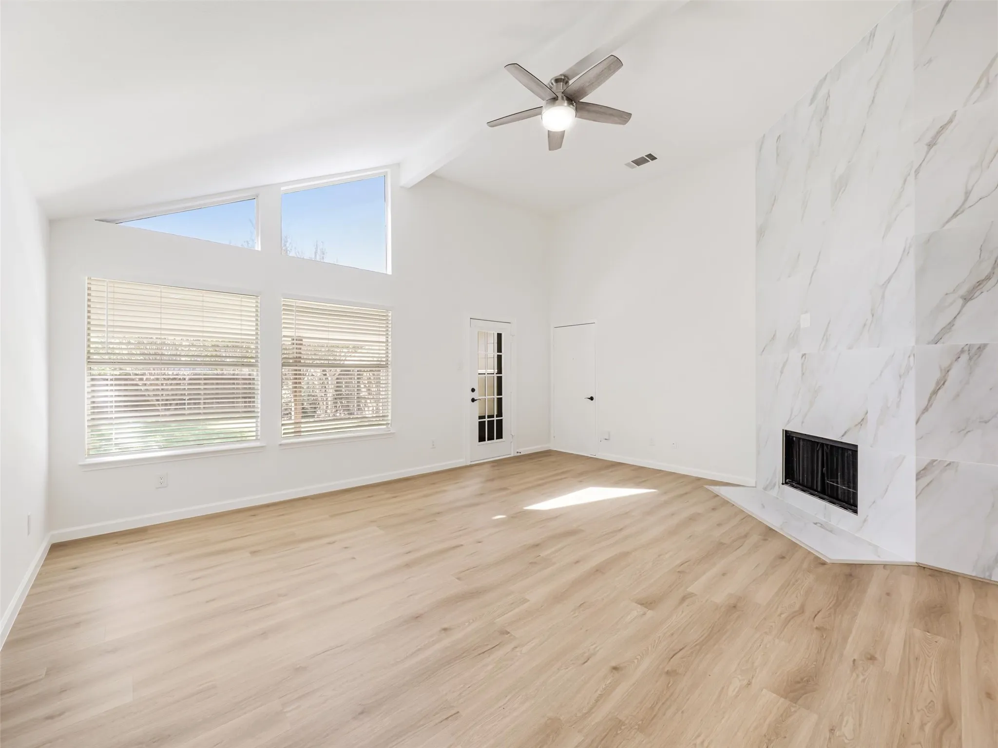 Vaulted ceiling with exposed beam, wood-finish flooring, white marble-look fireplace, large picture windows, and a ceiling fan