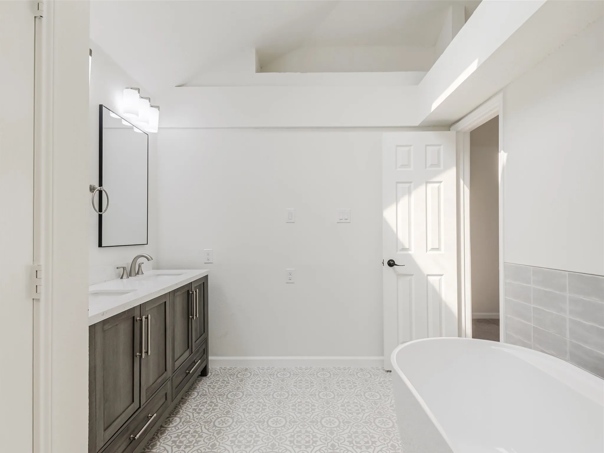 Dual vanity with undermount sinks, patterned floor tiling, freestanding soaking tub, and an accent wall with light gray rectangular tiles