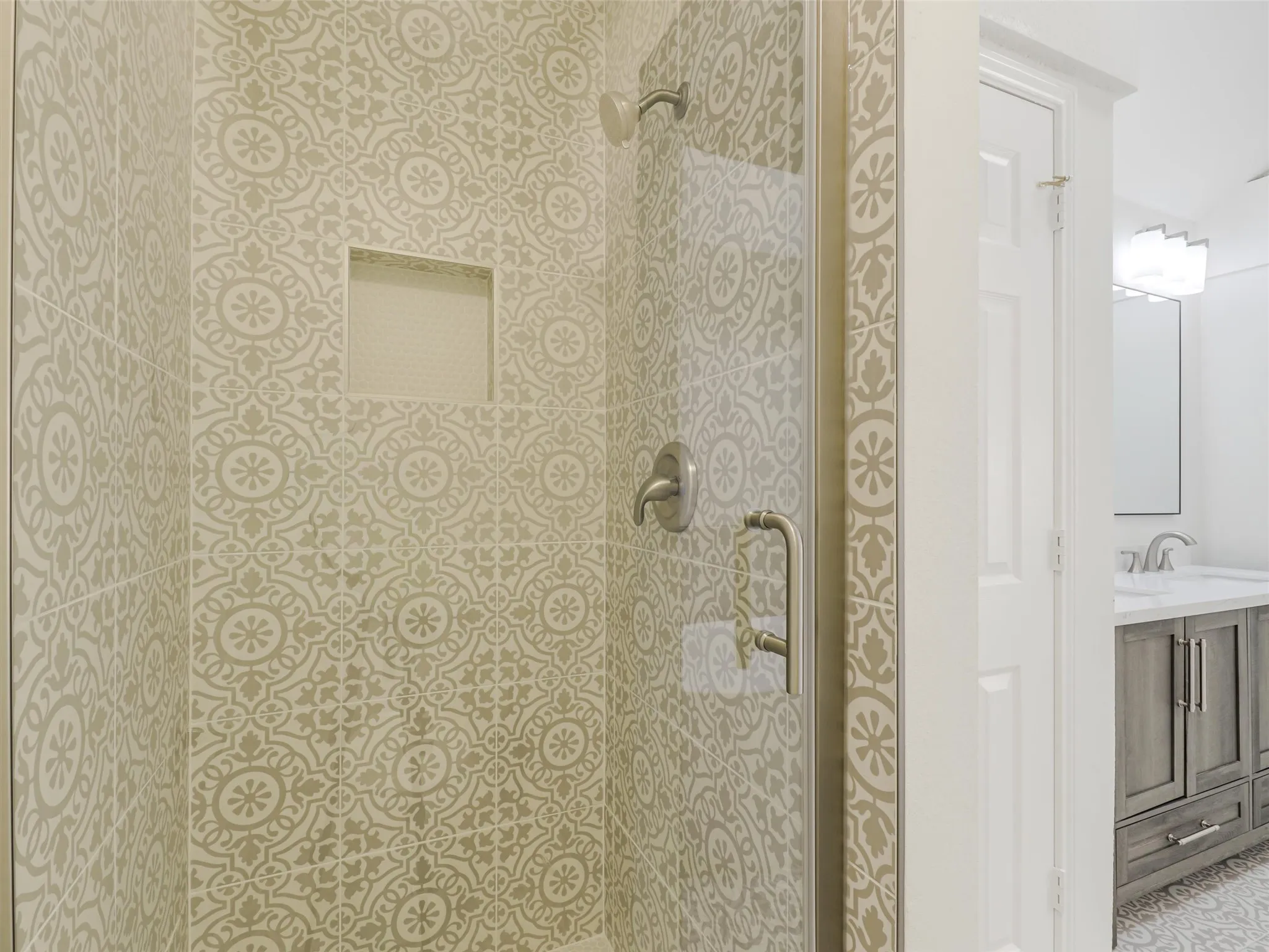 Patterned shower tile, built-in shower niche, brushed nickel fixtures, glass shower enclosure, and a vanity with a light-colored countertop