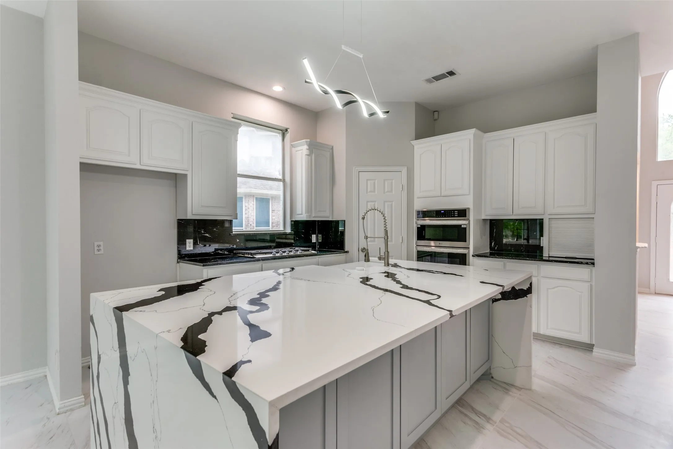 Spacious kitchen featuring a waterfall island with an integrated sink and modern faucet, white cabinetry, a built-in oven, and a contemporary linear chandelier