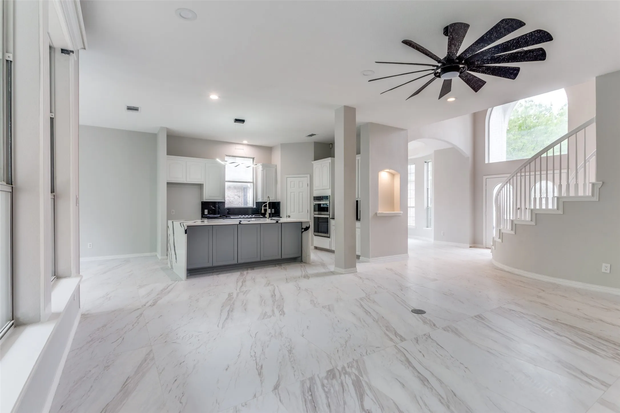 Expansive open-concept space featuring large format tile flooring, a kitchen island with stone waterfall countertop, built-in double ovens, and a grand curved staircase
