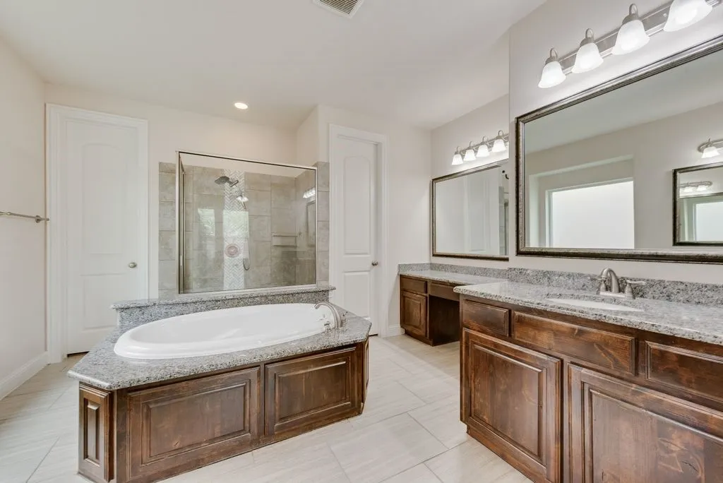 Spacious bathroom featuring an oval soaking tub with a granite surround, a glass-enclosed shower with tiled walls, dual wood-finish vanities with granite countertops, and large framed mirrors