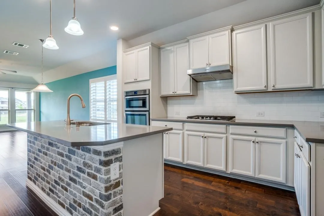Kitchen featuring white cabinetry, built-in double ovens, a gas range with range hood, a tiled backsplash, and a kitchen island with a brick finish and integrated sink