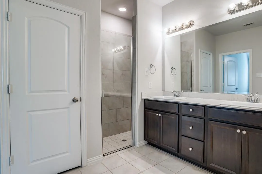 Dual vanity with dark wood-finish cabinetry and white countertops, large mirror, and glass-enclosed shower with tiled​​‌​​​​‌​​‌‌​‌‌​​​‌‌​‌​‌​‌​​​‌​​ walls
