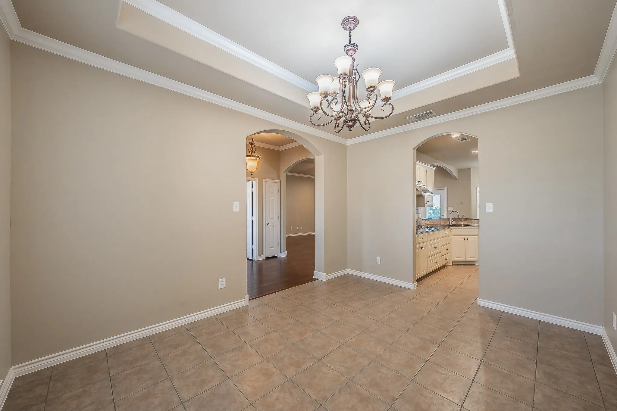 Tray ceiling with crown molding, decorative chandelier, tile flooring, arched doorways, and light-toned cabinetry