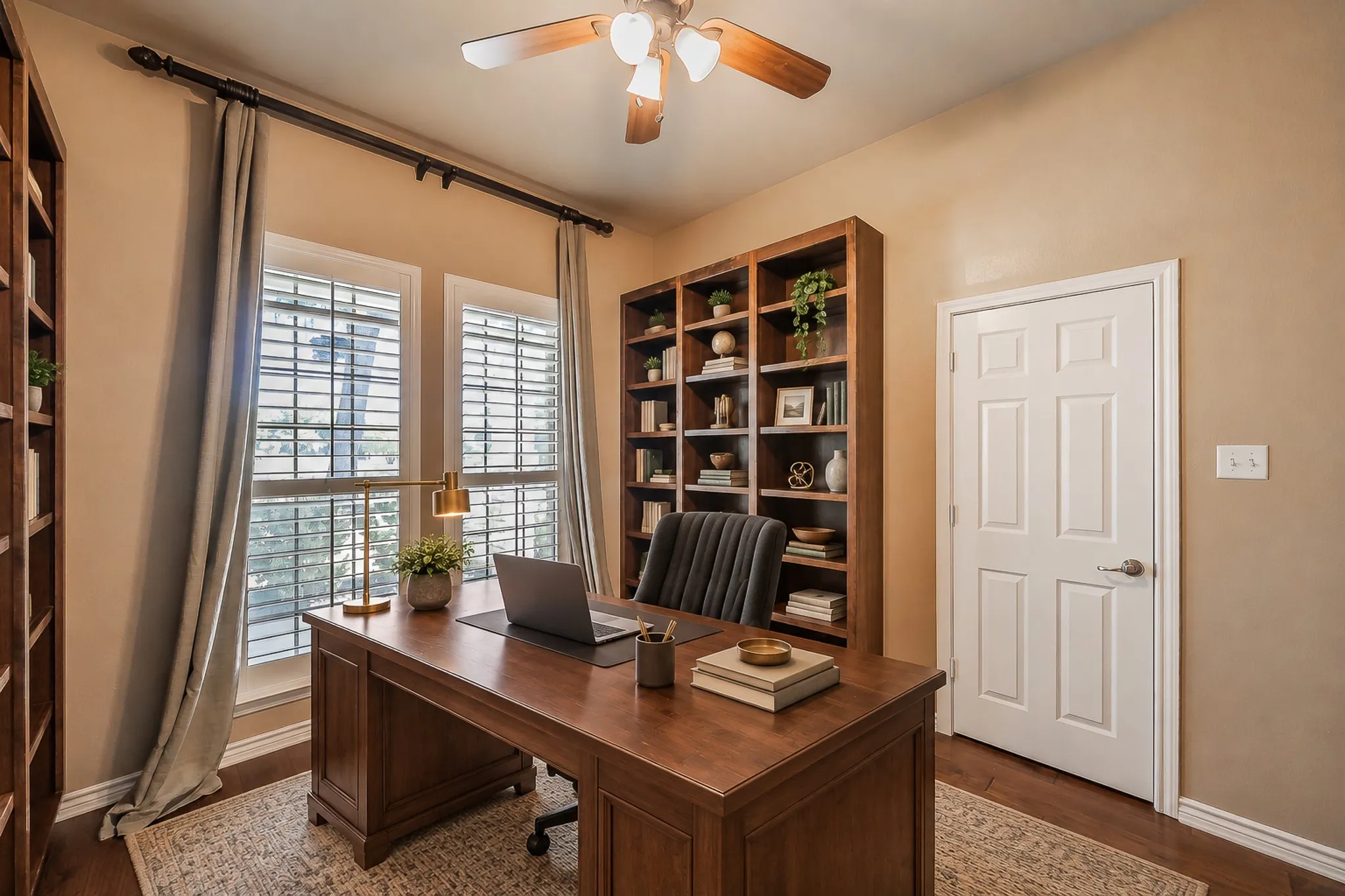 Rich wood-finish flooring, built-in shelving, two large windows with blinds, a ceiling fan, and a paneled white door