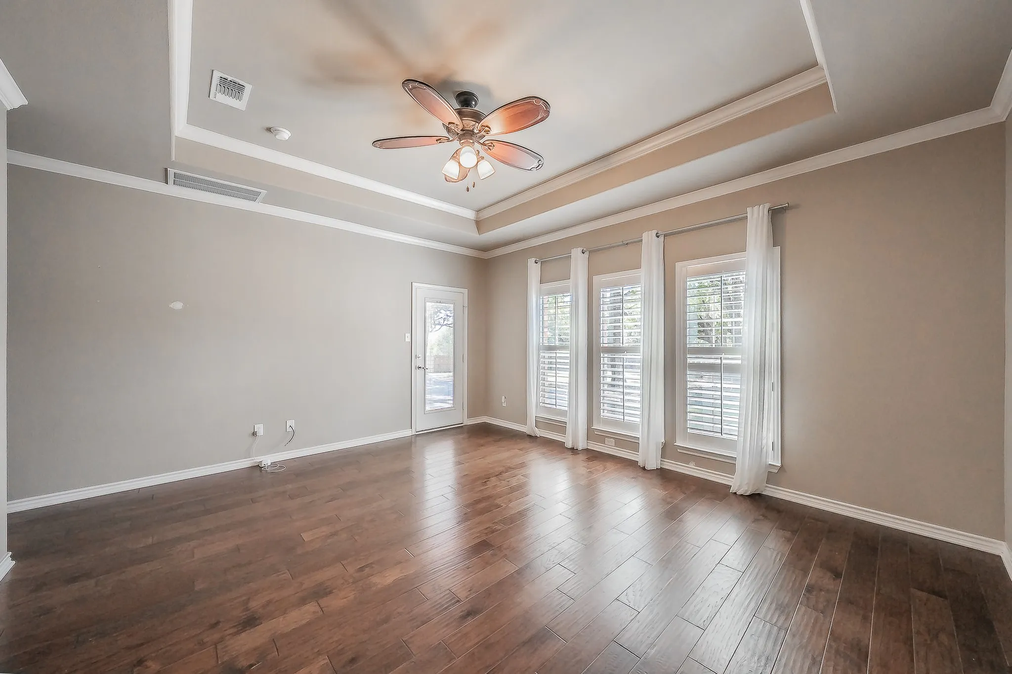 Wood-finish flooring, tray ceiling with crown molding, ceiling fan, three windows with plantation shutters, and a glass-paneled door