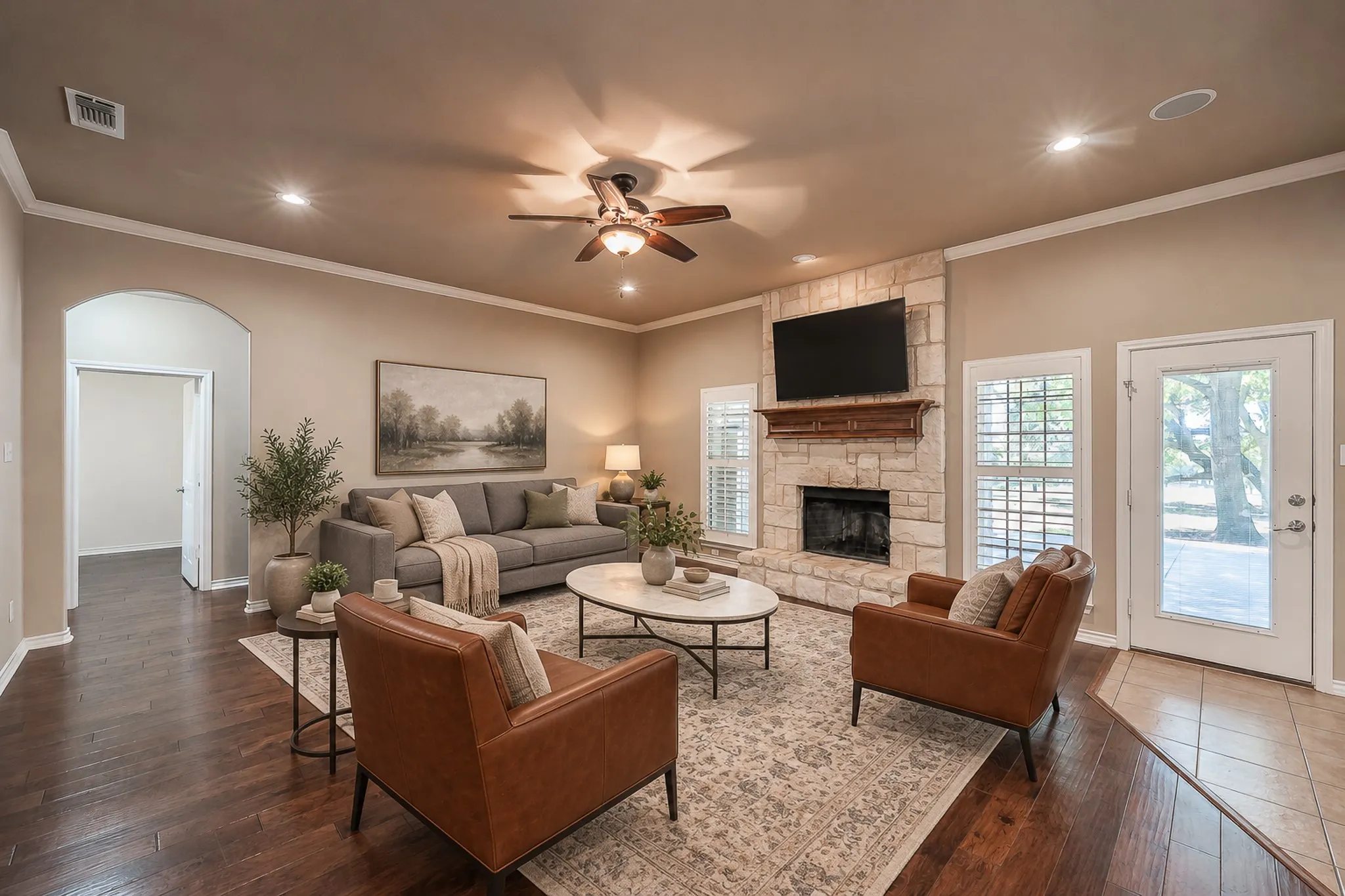 Spacious living area featuring wood-finish flooring, a stone-clad fireplace with wood mantel, crown molding, and a ceiling fan with integrated lighting