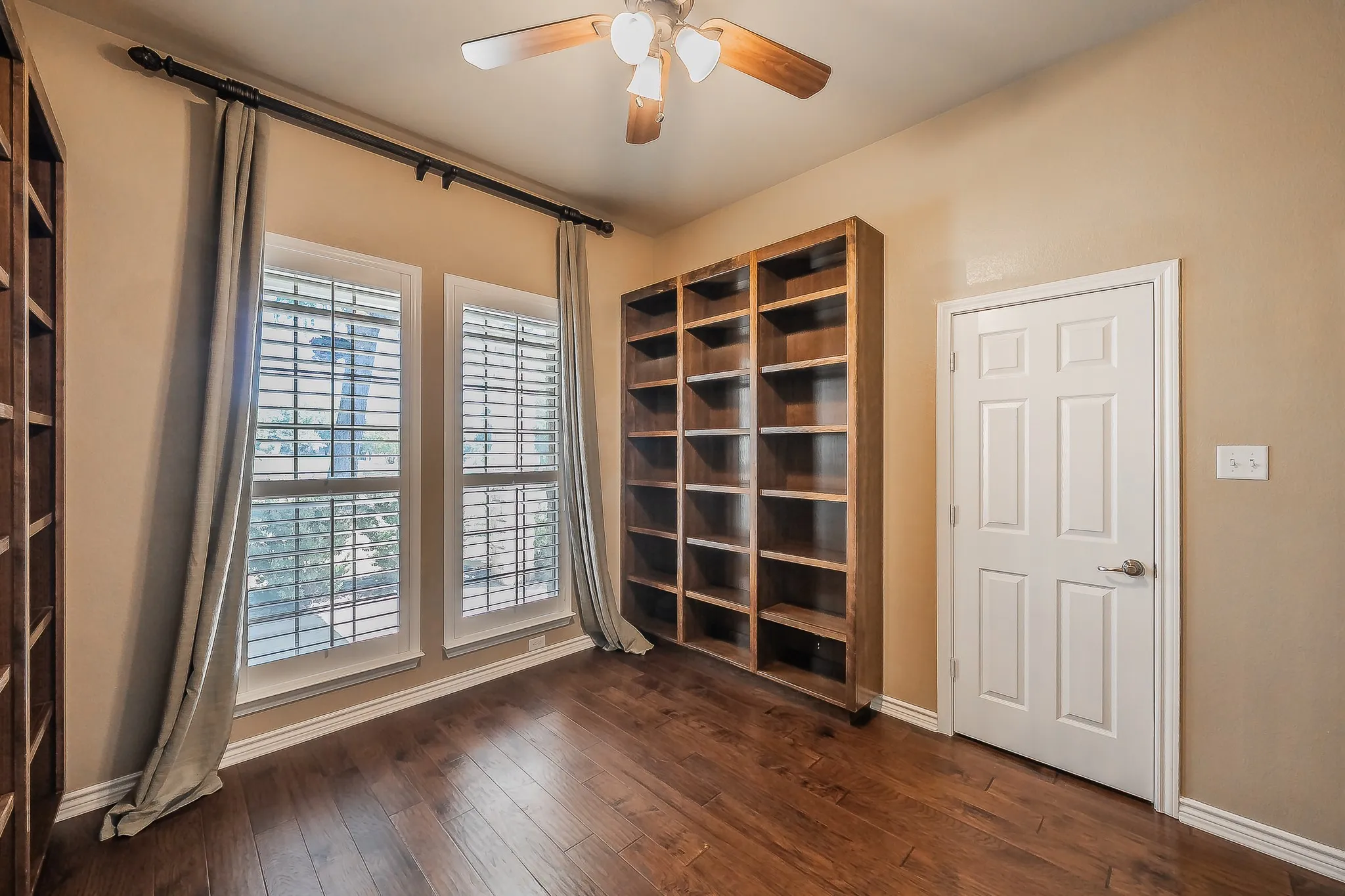 Wood-finish flooring, built-in shelving, two windows with plantation shutters, ceiling fan with integrated lighting, and a paneled door