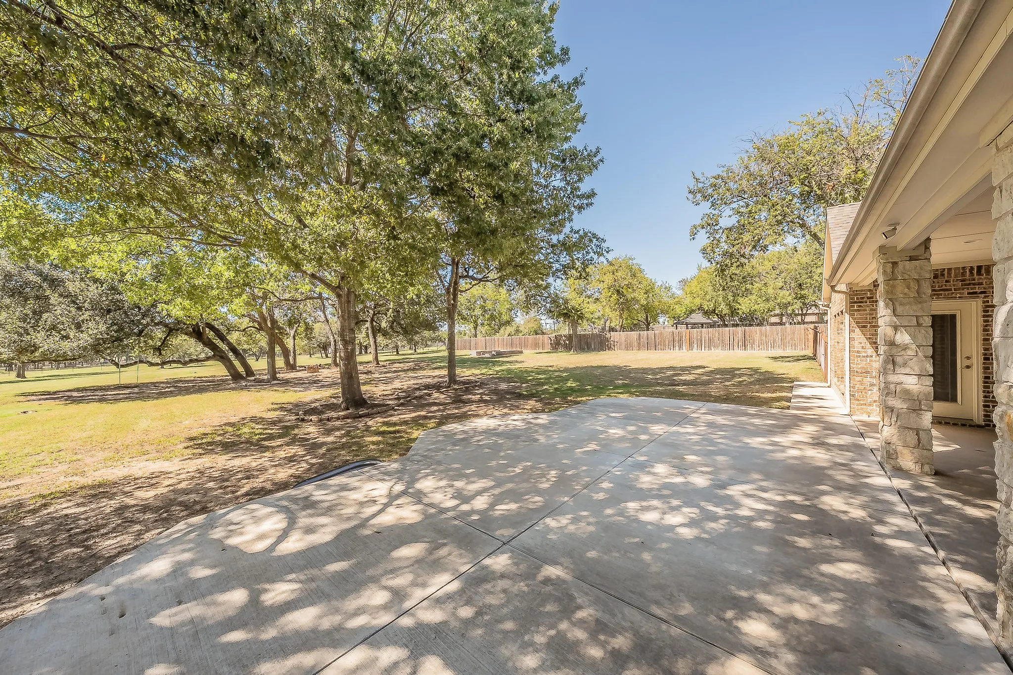 Expansive concrete patio with a partial stone and brick exterior, surrounded by mature trees and a manicured lawn