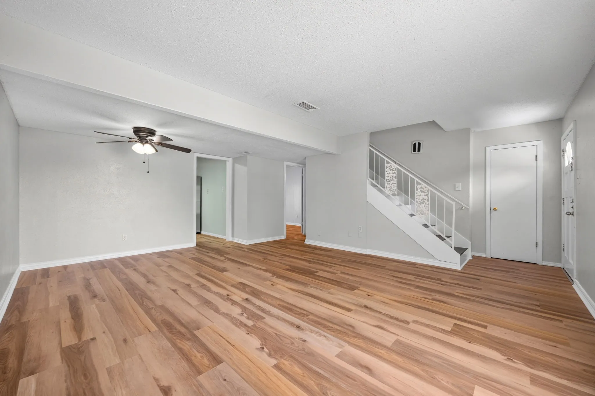 Spacious living area featuring wood-finish flooring, a ceiling fan with integrated lighting, and a prominent staircase with decorative railings